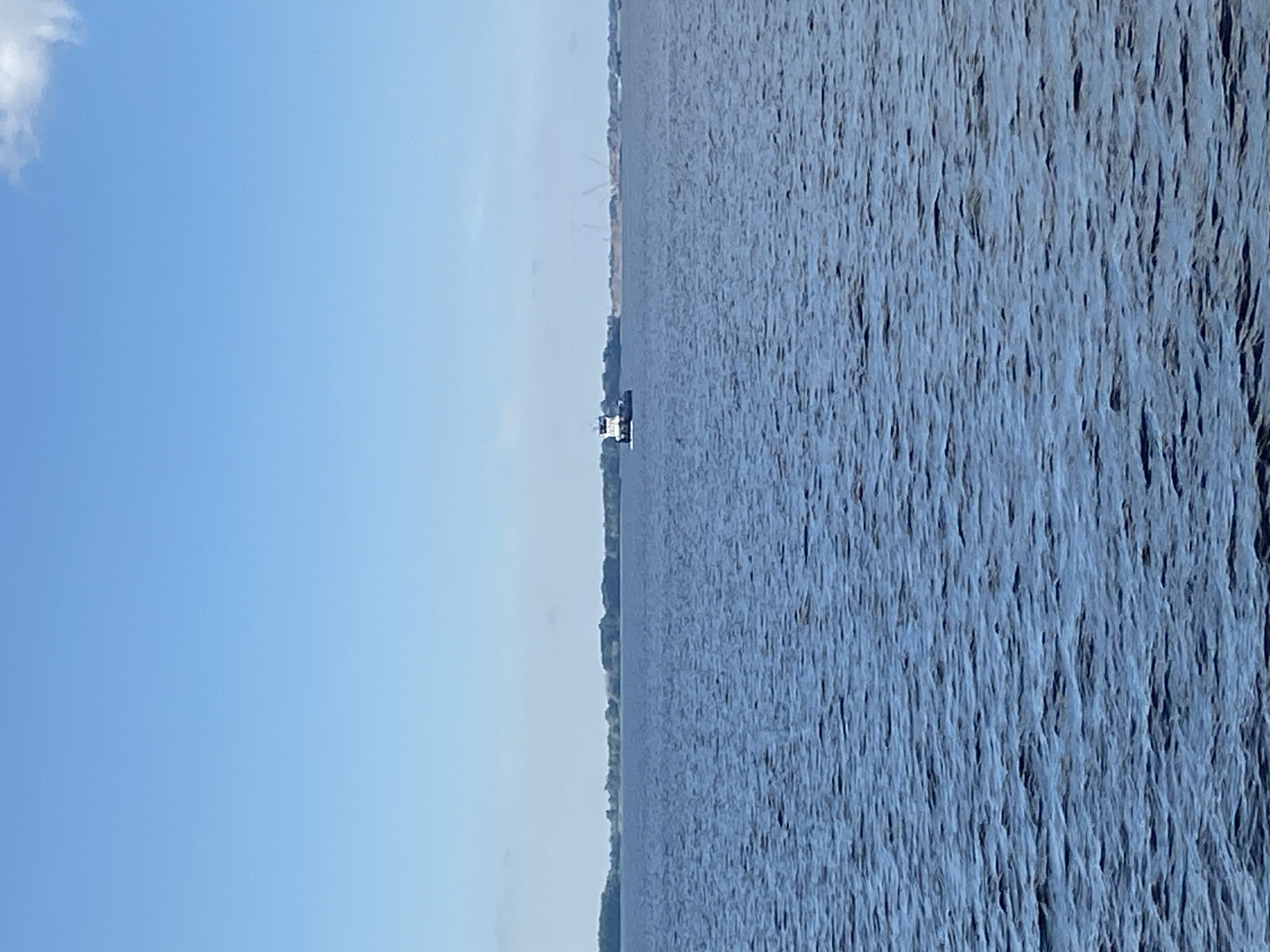 A tugboat is in the distance on a body of water under a clear blue sky. The tugboat is white with a black top and is moving away from the viewer, slightly to the left. The water is dark blue with small waves. In the background, a tree line is visible on the horizon, and in the far distance, there are cranes and structures that appear to be under construction. A small white cloud is in the upper right corner of the frame.