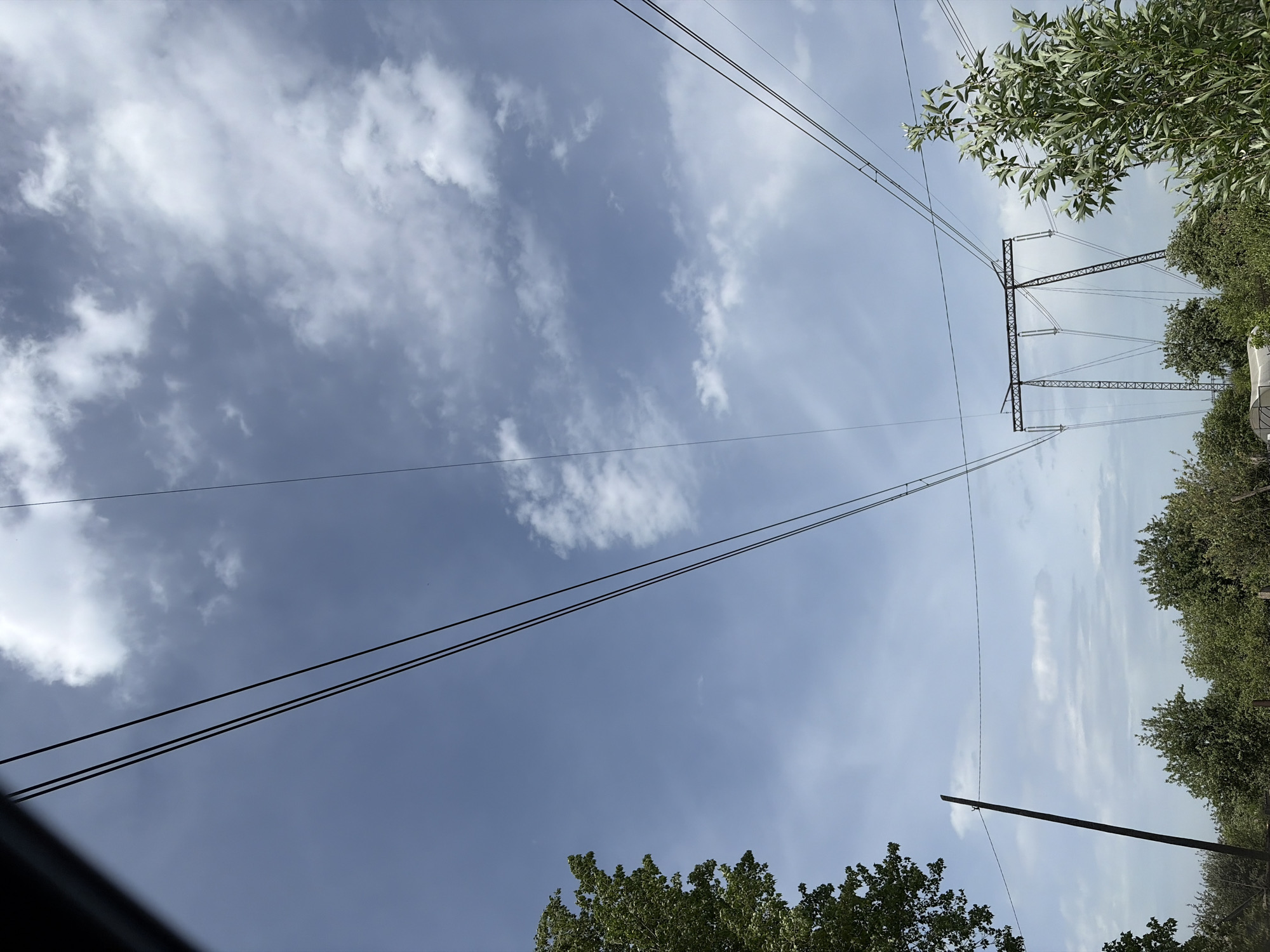 The image shows a view looking up at the sky with power lines and a power transmission tower visible. The sky is blue with scattered white clouds. The power lines run diagonally across the frame. The power transmission tower is visible on the right side of the frame, partially obscured by foliage. Green trees and bushes are seen along the edges of the frame. The location is likely Chaikovsky, Russia, as indicated in the prompt.