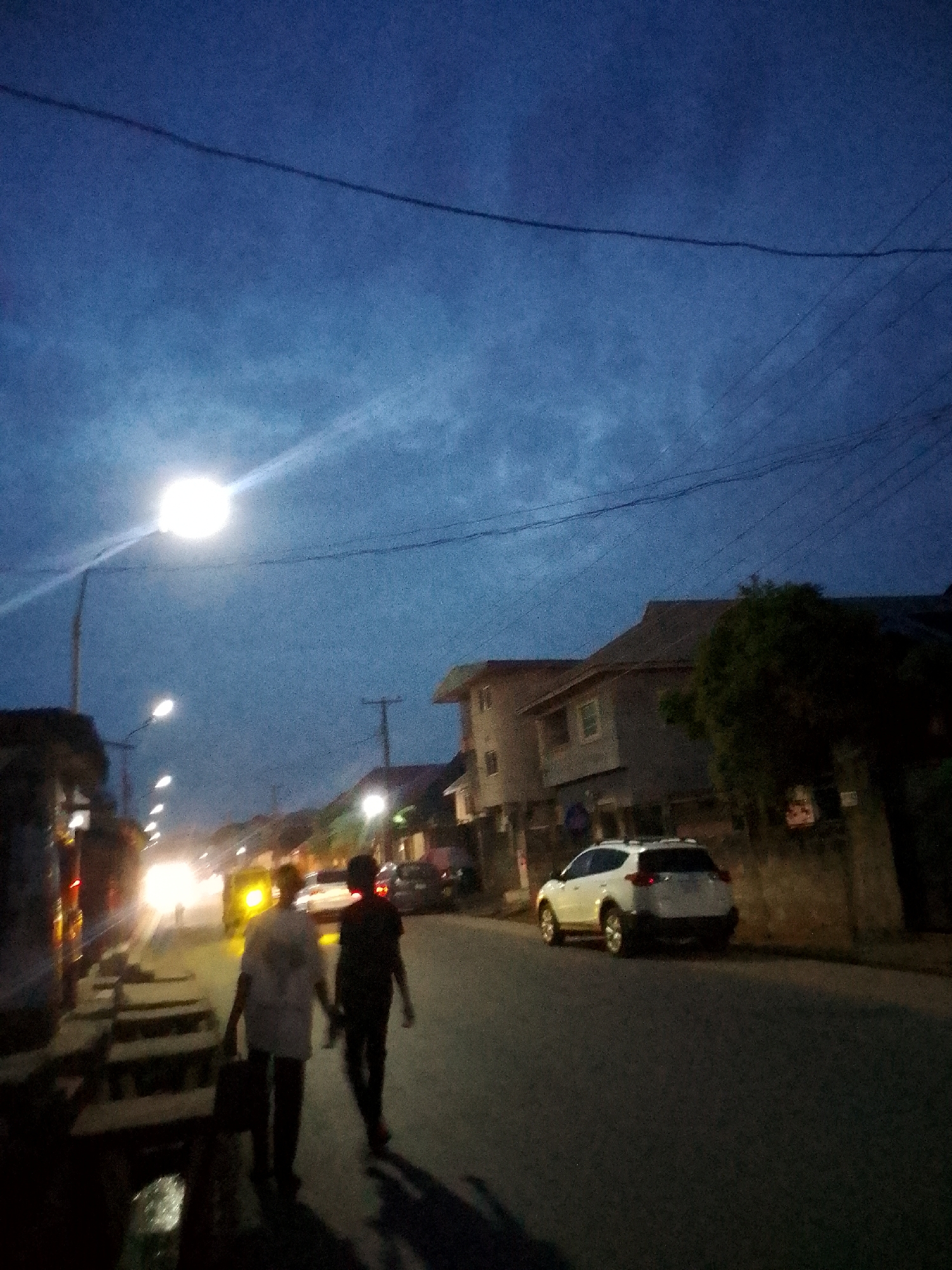The photograph shows a street scene in Onitsha, Nigeria, at dusk or night. Two individuals are walking along the road toward the camera. A light-colored car is parked on the right side of the road, near a building that appears to be a multi-story residential structure. Streetlights illuminate the scene, and there are other cars and buildings in the distance. Overhead power lines are visible against the dark sky. The overall light level is low.