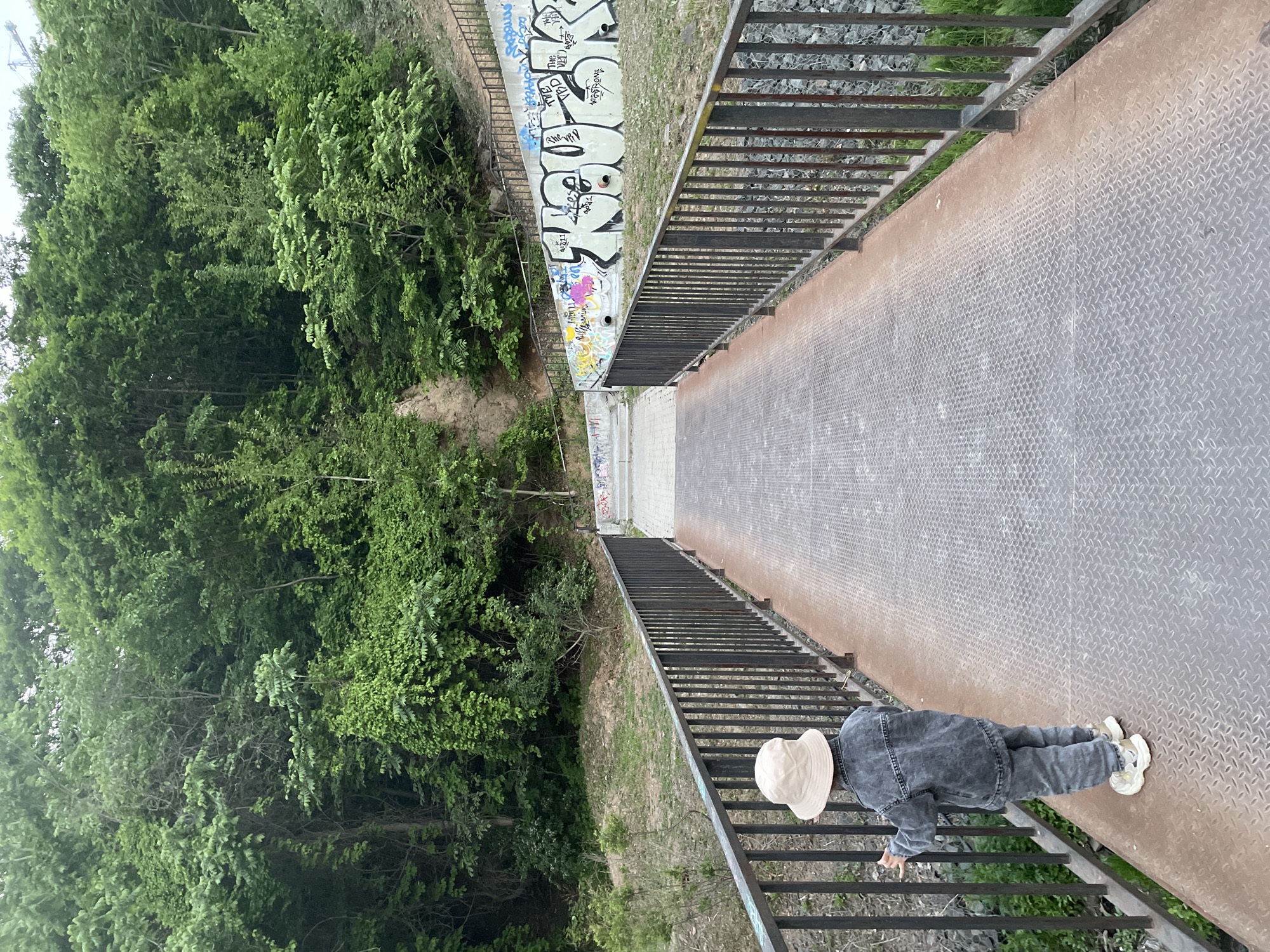 A sideways, high-angle shot shows a pedestrian bridge in Vladivostok, Russia. The bridge is constructed of steel with diamond plate flooring and dark-colored railing. The bridge spans a small ravine filled with trees and vegetation on the left. On the right, the wall alongside the ravine is covered in graffiti. A person wearing a light-colored hat, a denim jacket, and jeans is standing on the right side of the bridge, looking down into the ravine. The sky appears overcast.