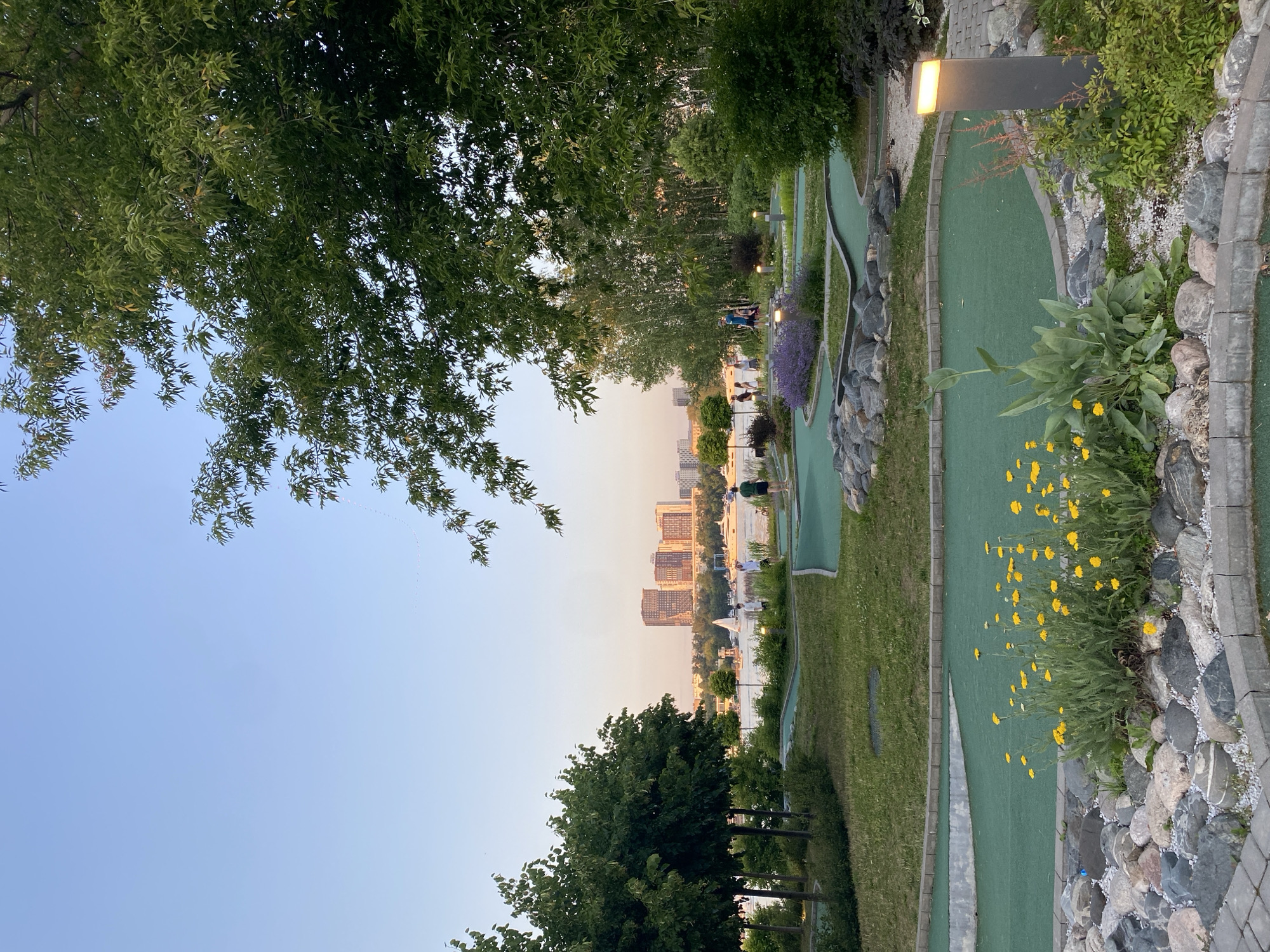 This is a medium shot, rotated image taken in Khimki, Russia. The scene depicts a miniature golf course adjacent to a waterfront with a city skyline in the distance. The time of day appears to be late afternoon or early evening, as indicated by the soft, warm lighting and the clear, pale blue sky.
The foreground is dominated by lush green trees on the left, with their branches reaching towards the sky. To the right, the miniature golf course is visible with its bright green turf, winding paths, and strategically placed rocks and landscaping. There are a few people scattered throughout the course, suggesting a recreational activity is in progress. Some individuals are seen walking or playing on the golf course, while others are gathered near the waterfront.
The middle ground features a body of water, likely a river or reservoir, with a few boats on it. The city skyline on the opposite bank is characterized by a cluster of multi-story buildings, suggesting an urban setting.
Notable details include the presence of ornamental plants, including vibrant yellow flowers and purple flowering bushes, adding color to the landscape. Streetlights are also visible along the path of the golf course, hinting at its use extending into the evening. The overall mood of the image is peaceful and serene, capturing a moment of leisure in an urban park setting. No visible text is present in the image.