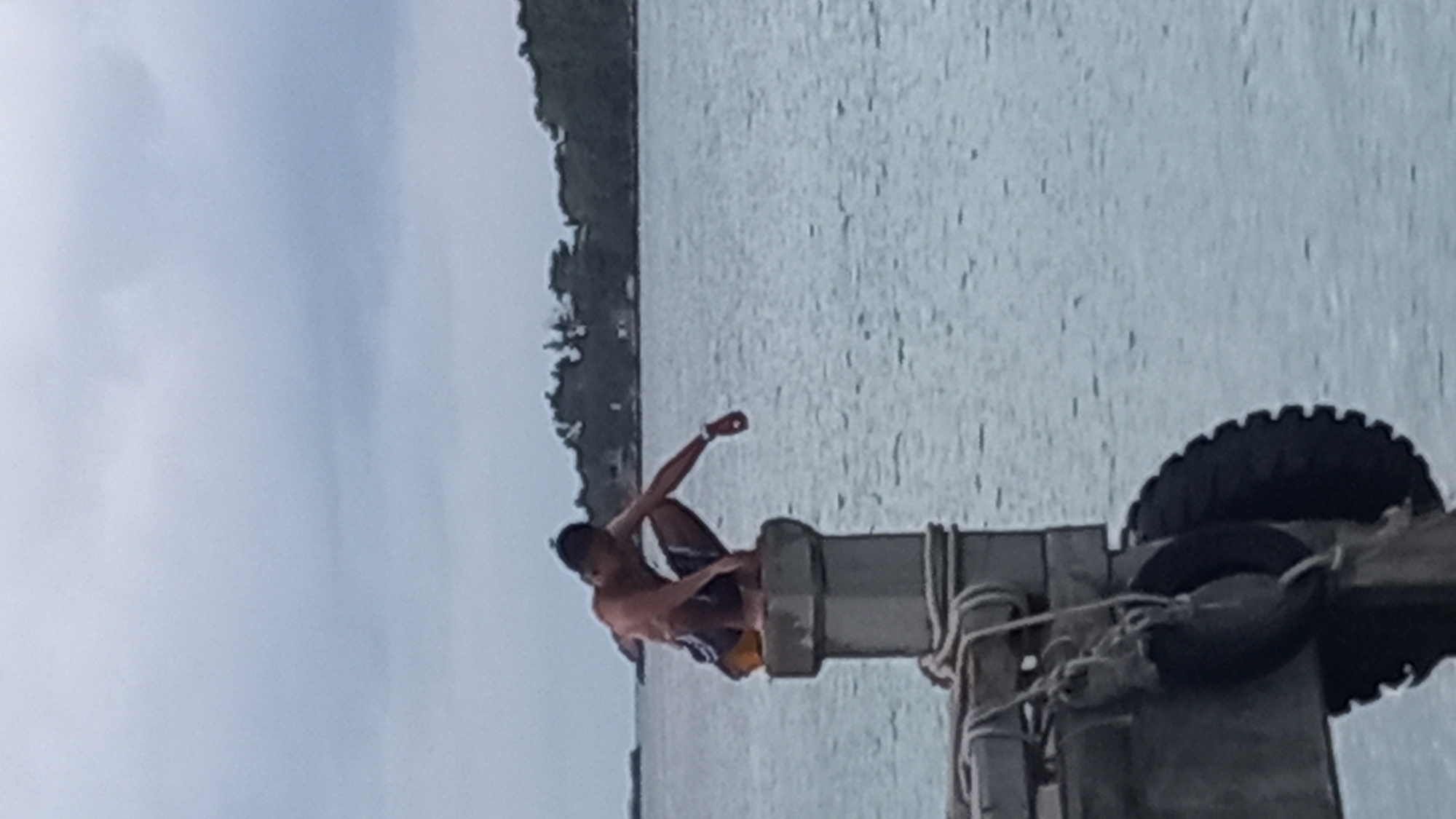 A young man is crouched on top of a concrete pillar on a pier, with his back to the camera and his head turned to the side. He is wearing dark-colored swim trunks with a yellow waistband. The water is choppy, and the sky is overcast. In the background, there is a line of trees on a distant shore. The pier structure is made of concrete and wood, with a tire hanging from a rope. The scene is located in Ko Pha-ngan District, Thailand.