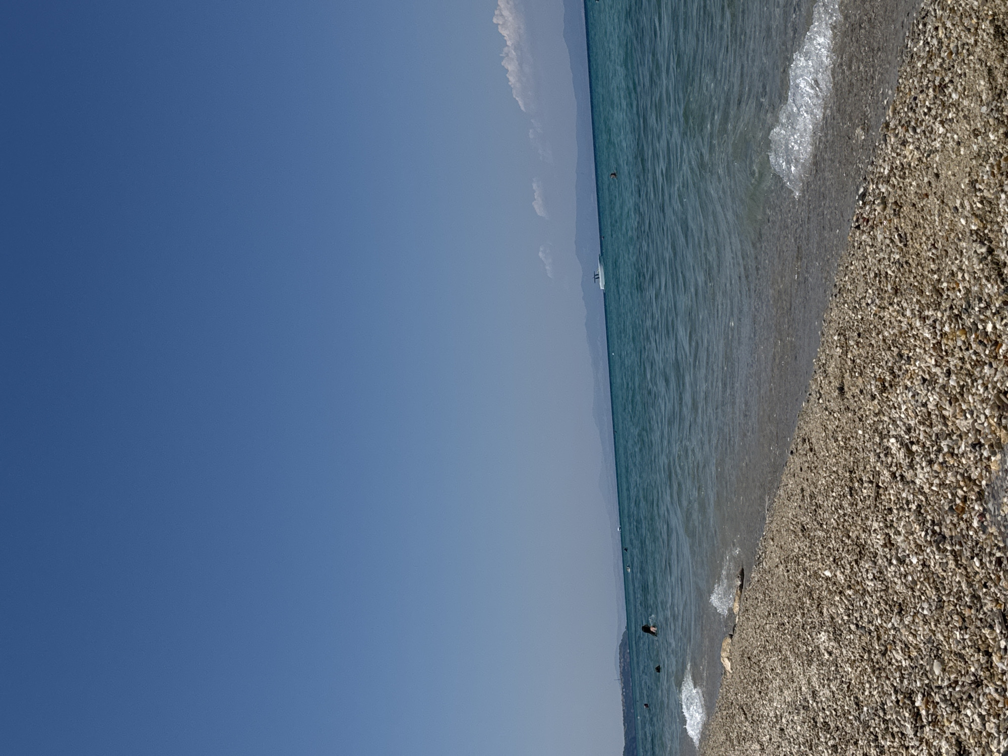 A wide-angle, eye-level shot captures a pebble beach with the turquoise waters of the Aegean Sea in Pallini Municipal Unit, Greece. The sky is a clear, cloudless blue. In the foreground, the beach is covered with numerous small, smooth pebbles of varying shades of brown, grey, and white. Gentle waves lap at the shore, creating a thin line of foam. Further out in the water, several people are visible swimming or wading. A white motorboat is sailing in the distance, and a faint coastline with mountains is visible on the horizon.