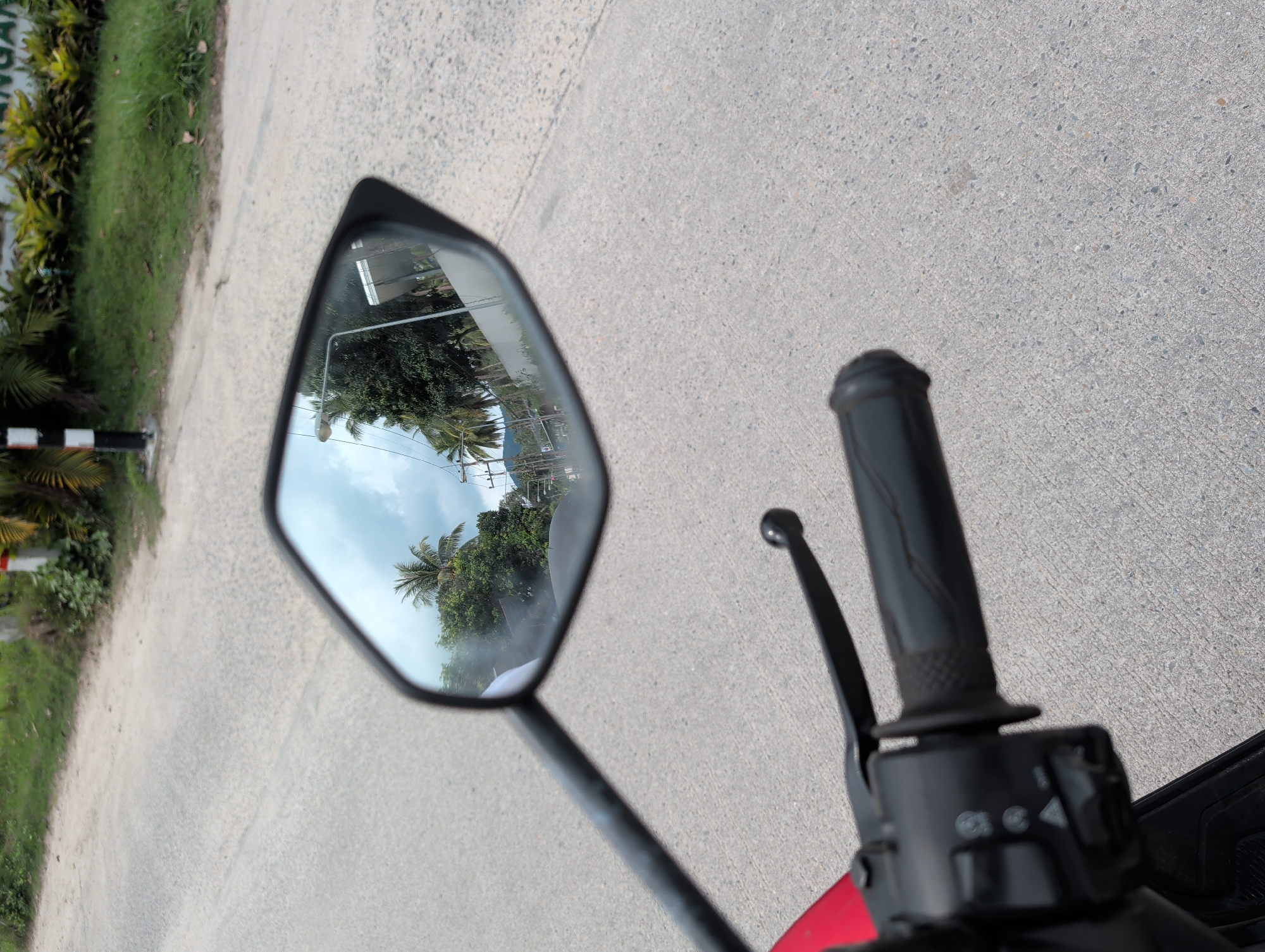 The image is taken from the perspective of a rider on a motorcycle traveling on a paved road. The rider's right handgrip and rearview mirror are visible. The rearview mirror reflects a scene of a road lined with trees and utility poles. The reflected image appears to be a road in a populated area with residential or commercial buildings. The road surface is a light gray concrete. The environment suggests a tropical or subtropical climate due to the presence of palm trees. Based on the description, the location is identified as Moo 4, Thailand.