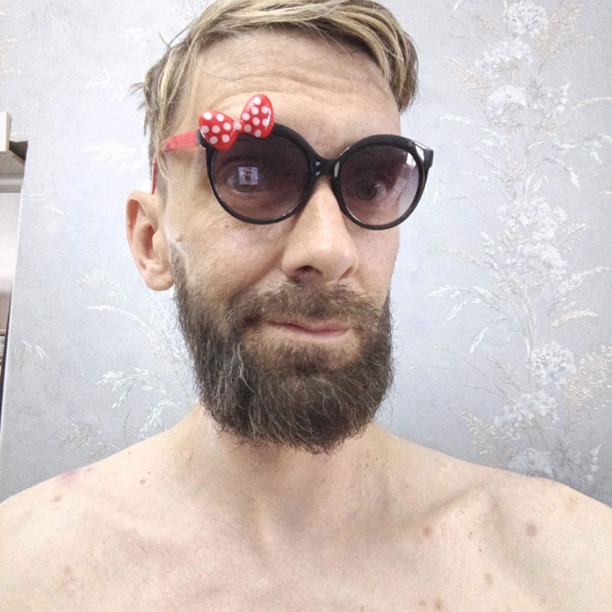 A Caucasian man with a blond, partially disheveled haircut and a beard is facing the camera, wearing black sunglasses with a red and white polka-dotted bow attached to the frame. He is bare-chested. The background is a wall with a floral pattern. The location is Марковский, Russia.