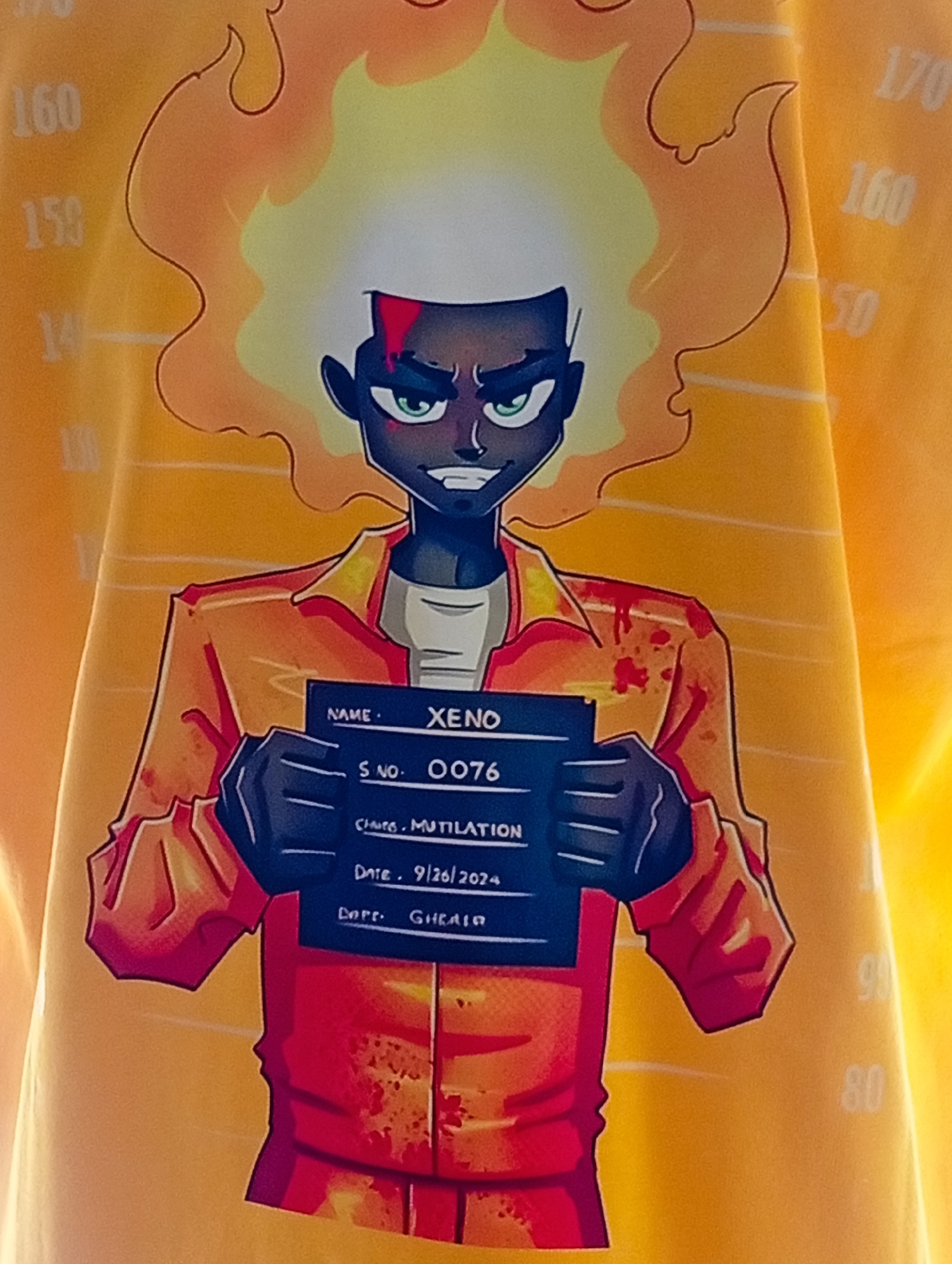 The image is a close-up shot of a yellow t-shirt with an illustration of a character holding a mugshot sign. The character is a dark-skinned person with bright green eyes and fiery orange-yellow hair. They are wearing an orange prison jumpsuit with red splatters, suggesting bloodstains. The mugshot sign is dark blue and contains handwritten text, including "NAME: XENO," "S NO. 0076," "CHARGE: MUTILATION," "DATE: 9/26/2024," and "CPFF: GHEAIR." Vertical lines with numbers are visible on the left and right sides of the image, resembling a height chart found in a police station. The background of the t-shirt is a solid yellow, with some horizontal lines indicating a fabric texture. The image does not depict any location in the city of Bukuru, Nigeria.