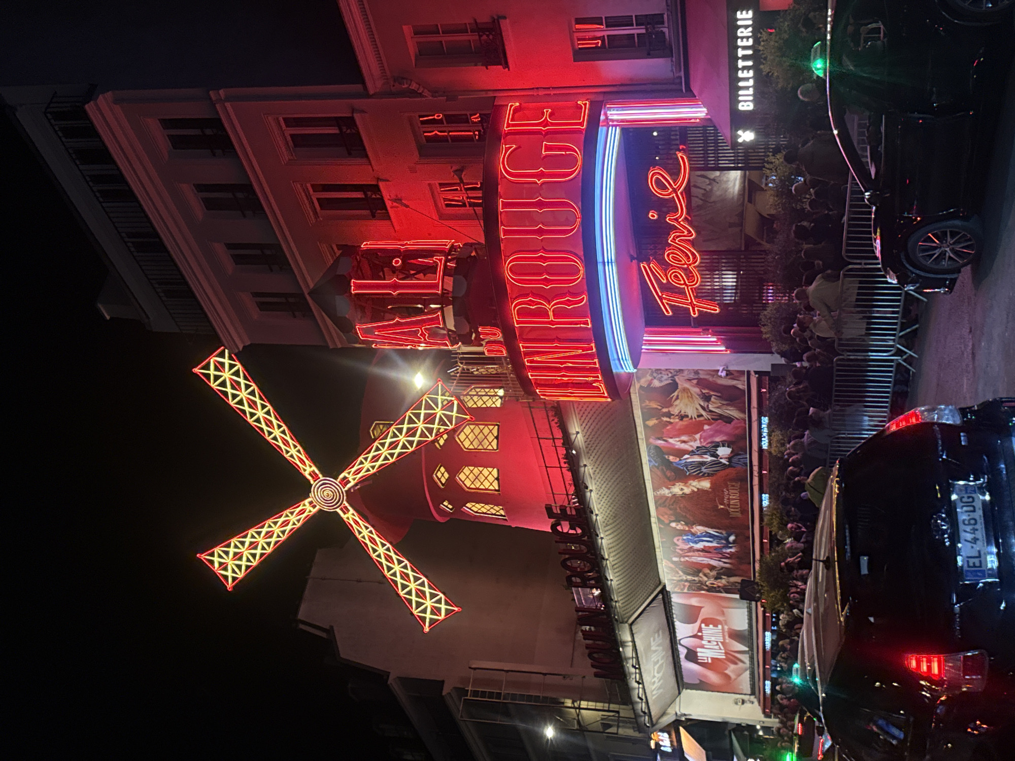 The image portrays the Moulin Rouge, a cabaret in Paris, France, at night. The iconic red windmill atop the building is lit with glowing yellow and red lights. The building's facade is also illuminated in red, and neon signs spell out "Moulin Rouge" and "Féerie" in blue and red. A crowd of people stands outside the entrance, possibly waiting to enter or watch a show. Advertisements and posters flank the entrance and a black vehicle is parked on the right side of the image, a number plate partially visible "EL-446-DG". The overall setting suggests a lively atmosphere typical of Parisian nightlife.