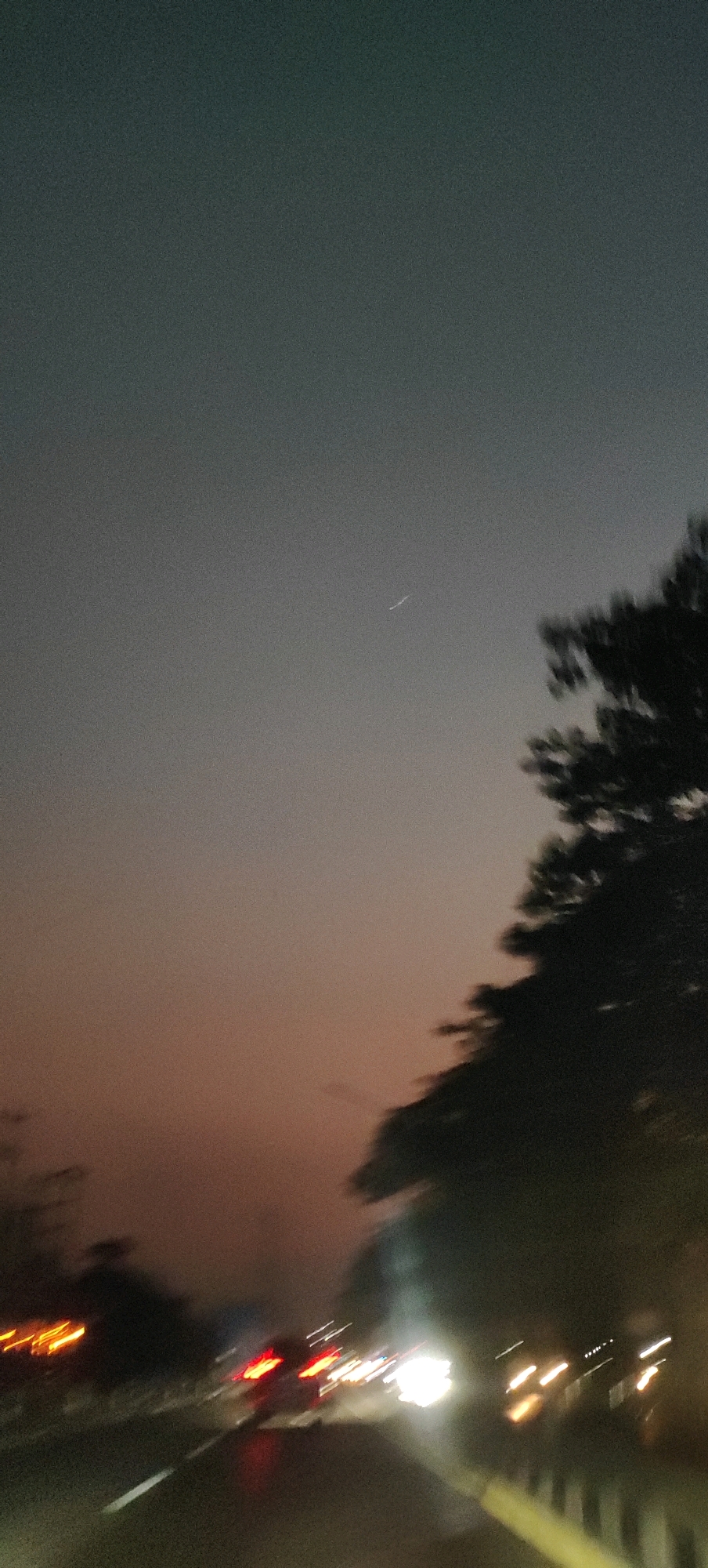 The image depicts a blurry, nighttime scene from a vehicle traveling on a road. The sky is dark with a gradient of colors from a muted grey at the top to a reddish-orange near the horizon. A faint white streak, possibly a distant airplane or meteor, is visible in the upper sky. On the right side of the frame, the silhouette of a tree with blurred leaves is visible. The foreground shows a road with white lane markings and the blurred lights of other vehicles. Red taillights and white headlights streak across the scene, indicating movement. The overall impression is one of a fast-moving perspective on a road at dusk or night, with the ambient light creating streaks and blurs. The location is identified as Sedo Lor, Indonesia.