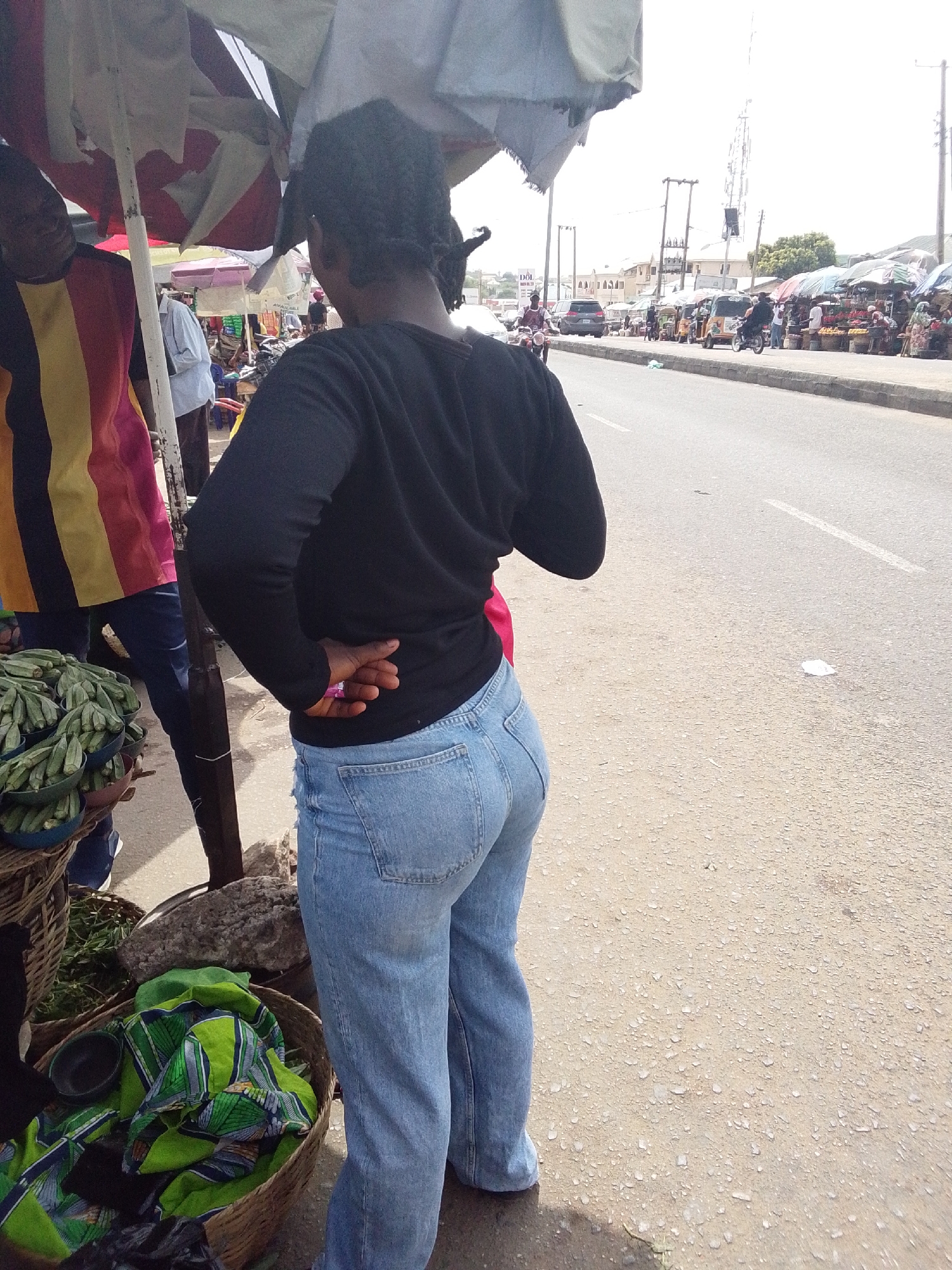 A woman wearing blue jeans and a black top stands in Adankolo, Nigeria, near a roadside market. She faces away from the viewer, her hands clasped behind her back. Beside her are baskets of okra and a basket of green and blue patterned fabric. A man in a striped shirt stands to her left under a white and red striped umbrella. The road stretches ahead with various vehicles and market stalls visible in the distance.
