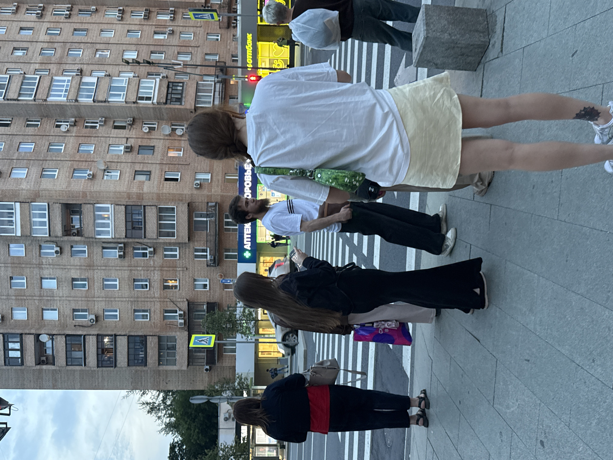 The image depicts a street scene in Moscow, Russia, likely during the late afternoon or early evening, given the ambient light. In the background, a multi-story brick apartment building dominates the frame, featuring numerous windows and air conditioning units. A street with a pedestrian crossing is in the foreground, with several individuals visible.
On the left, a woman with dark hair, wearing a dark jacket over a red shirt and dark pants, stands with her back to the camera, holding a light-colored handbag. Next to her, another woman with long, dark hair, dressed in a black top and flared black pants, is also seen from behind, holding a smartphone and a colorful shopping bag. A man with a beard and a white t-shirt is standing to her right. In the center of the frame, a person wearing a white t-shirt and a light-colored skirt is facing away, carrying a green patterned bag. To the far right, another individual in dark pants and a dark top is seen from the back, holding a large white plastic bag.
Across the street, there are storefronts with illuminated signs, one of which displays Cyrillic text. A silver car is visible on the road. The pedestrian crossing is marked with white stripes. The sidewalk is paved with large, rectangular gray tiles. A gray concrete bollard is present on the right side of the sidewalk. Traffic lights are visible, indicating a street intersection. The sky above is overcast with some light visible. The overall atmosphere suggests an urban setting with people engaged in everyday activities.