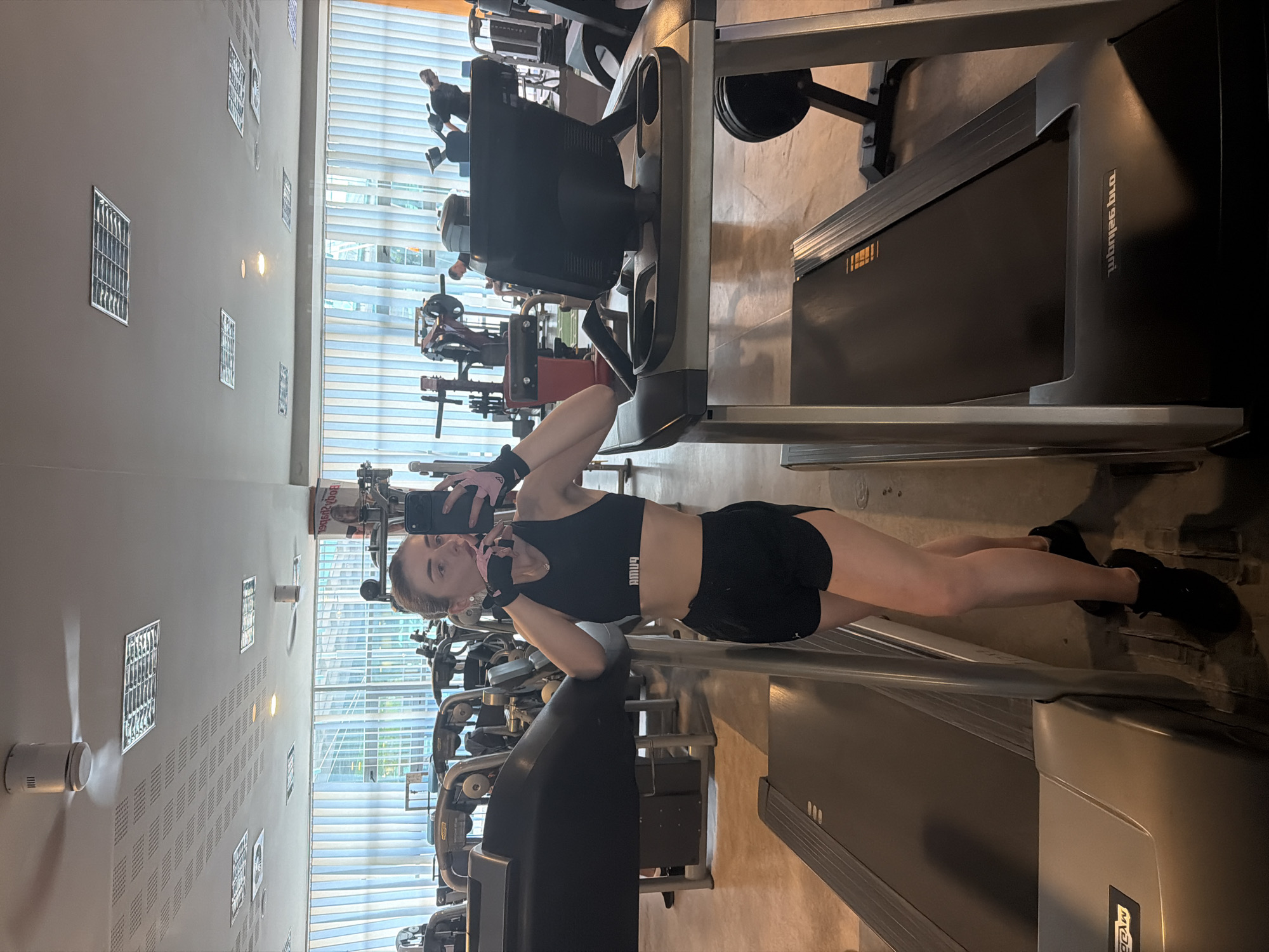 A young woman with her hair pulled back stands in a gym, taking a selfie in a mirror. She is wearing a black Puma sports bra and black athletic shorts, along with black sneakers and fingerless gloves. She is leaning on a treadmill, and another treadmill is visible to her left. The gym has large windows with vertical blinds, and fluorescent lighting is visible on the ceiling along with two ceiling fans. Other gym equipment, including weight machines, can be seen in the background. The floor is a light brown or tan color, and the overall environment appears to be an indoor fitness facility.