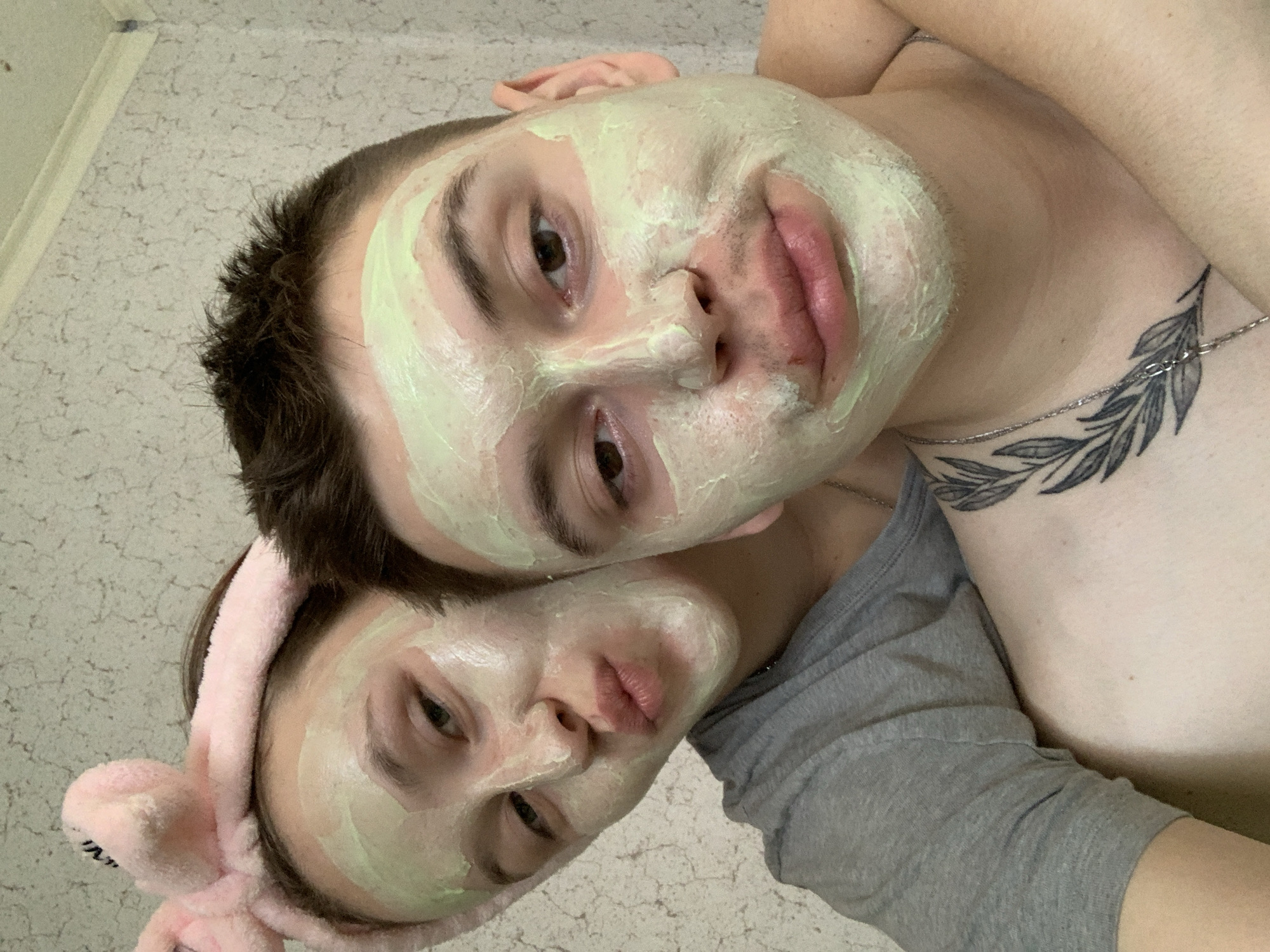 This close-up, tilted selfie captures two individuals, likely a couple, engaged in a shared skincare routine in an indoor setting. Both subjects have a light green facial mask applied generously to their faces.
The person in the foreground, positioned lower and to the left, appears to be female. She wears a soft, pink fuzzy headband, possibly with bunny ears, keeping her hair away from her face. Her eyes are open and directed towards the camera, with her lips slightly puckered. She is partially visible wearing a grey t-shirt.
Behind and slightly above her is a male individual, also looking into the camera with open eyes and a subtle pout. A silver chain necklace adorns his neck. A prominent tattoo depicting leafy branches is visible on his upper chest and shoulder area. He appears to be shirtless, or wearing clothing that exposes his upper torso.
The image is taken from a high angle, looking down at the subjects who are positioned close together. The background is a light-colored, textured surface, possibly a tiled wall or ceiling, suggesting a casual home environment, potentially in Фоки, Russia. The overall atmosphere is intimate and relaxed, conveying a sense of shared, playful self-care. There is no visible text in the image.