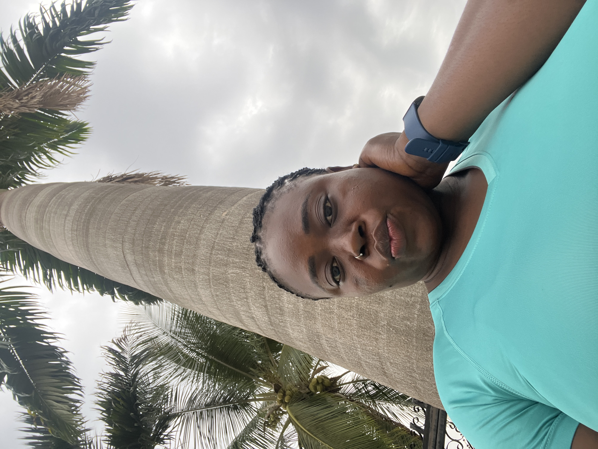 A woman is taking a selfie in front of a large palm tree, with more palm trees and a cloudy sky in the background. The woman has dark skin, braided hair, and is wearing a turquoise t-shirt and a dark blue smartwatch. She has a nose ring and her hand is touching her neck. The image appears to have been taken outdoors in a tropical or subtropical environment. Based on the context, the location is the city of Abuja, Nigeria.