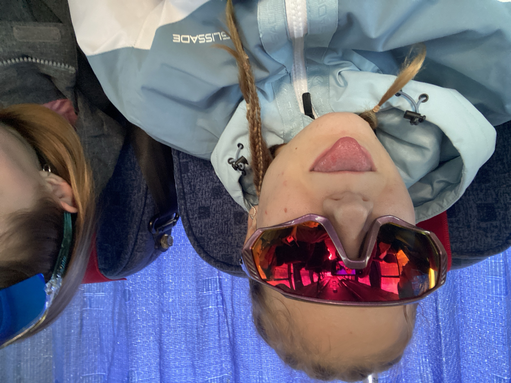 Two individuals are depicted in this photograph. The primary subject is a person wearing reddish-mirrored ski goggles and a light blue ski jacket with the word "BLISSADE" on it. They are sticking their tongue out. The person has fair skin and braided hair. A second individual is partially visible on the left side of the frame. This person is also wearing ski goggles. The background appears to be a blue textured surface. There is insufficient information to confirm or deny any details related to Berezniki, Russia.