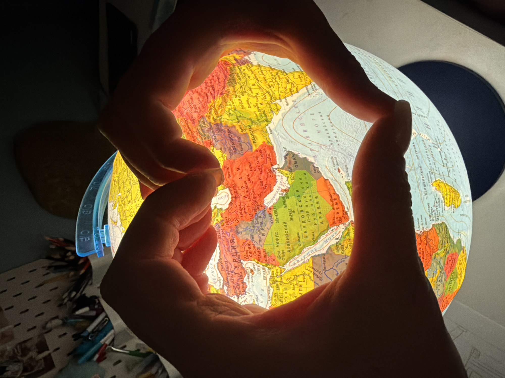 A pair of hands forms a frame around a backlit globe, highlighting the Middle East and Central Asia. The globe's map is detailed, with labeled cities and geographical features in Cyrillic script. The hands obscure the edges of the globe, creating a bright area in the center and shadows along the perimeters. A dark background indicates the photo was taken indoors. Other objects, including what seems like a desk and a pen holder, are partially visible. The location is not apparent from the image.