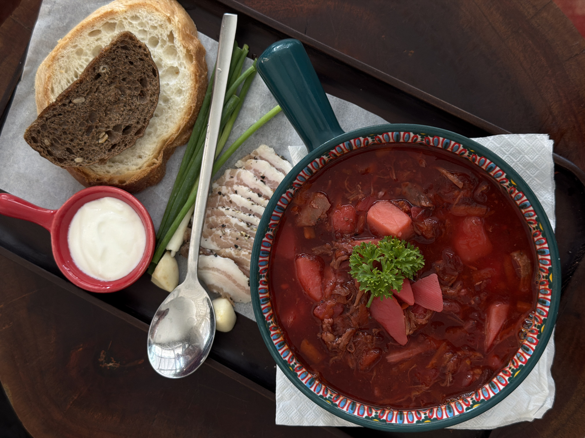 The image presents a tray with a serving of borscht and accompanying elements, situated in what appears to be Đà Nẵng, Vietnam. The main subject is a bowl of borscht, a beet-based soup, garnished with parsley and possibly pickled elements, served on a patterned bowl with a green handle. A white linen napkin supports the bowl. Adjacent to the bowl, there's a serving of bread with two slices – one light and one dark – next to a small red bowl of sour cream. Raw spring onions and sliced pork belly (possibly Lardo), and garlic cloves are also present. A silver spoon is placed beside the bread and sour cream. The tray is made of dark wood. No people are visible in the image. The scene suggests a meal setting in a Vietnamese location.