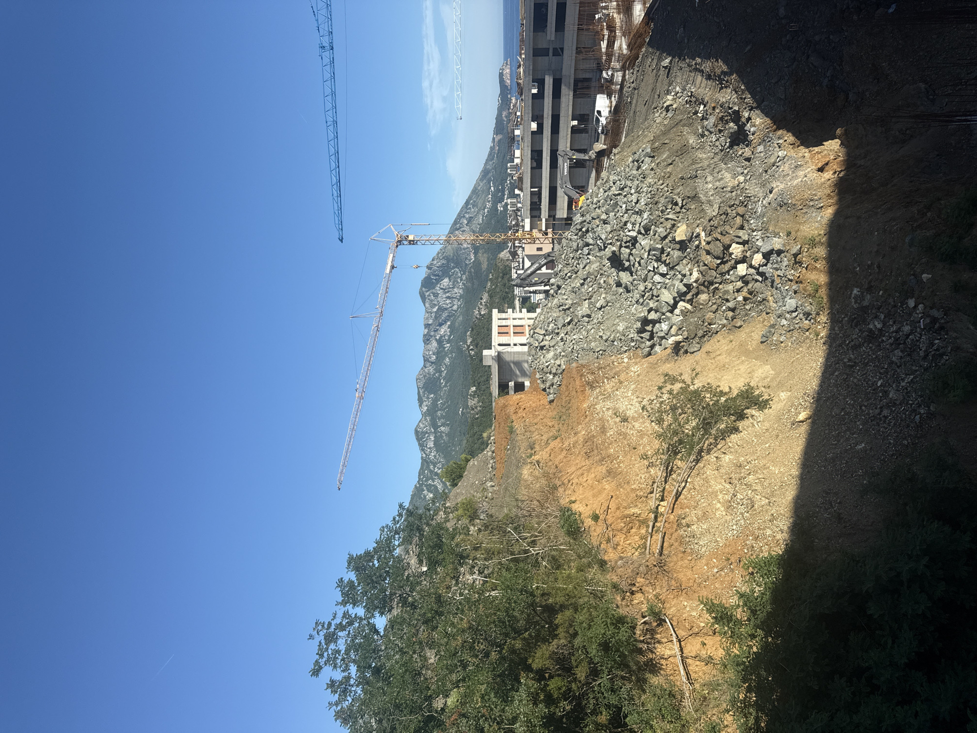 The image depicts a construction site in Boreti, Montenegro, on a clear day. A large, unfinished concrete building structure is visible in the middle ground, with several construction cranes, including a yellow tower crane and a blue mobile crane, positioned around it. The site appears to be on a hillside overlooking the Adriatic Sea, with mountains in the background. A large pile of rubble and excavated earth dominates the foreground, with vegetation on the left side of the image. The scene is devoid of any people.