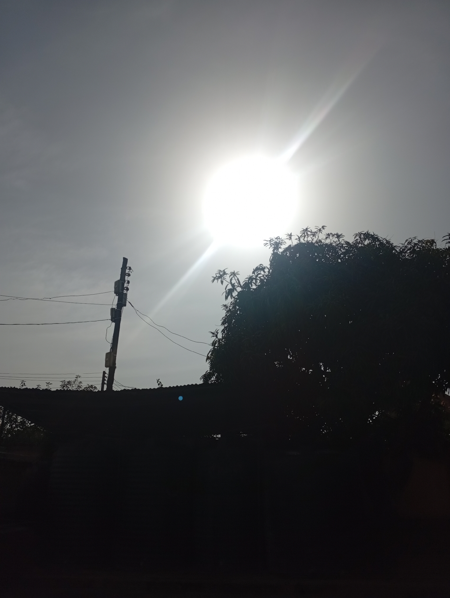 The image depicts a bright, overcast sky with the sun prominently featured, casting a strong glare. In the foreground, silhouetted against the bright sky, is a utility pole with electrical equipment, power lines stretching across the frame, and a corrugated metal roof structure. A dense tree canopy with visible leaves is also silhouetted on the right side of the image. The overall scene suggests an outdoor setting, possibly in a residential or semi-urban area. The location is identified as Rafin Par, Nigeria.