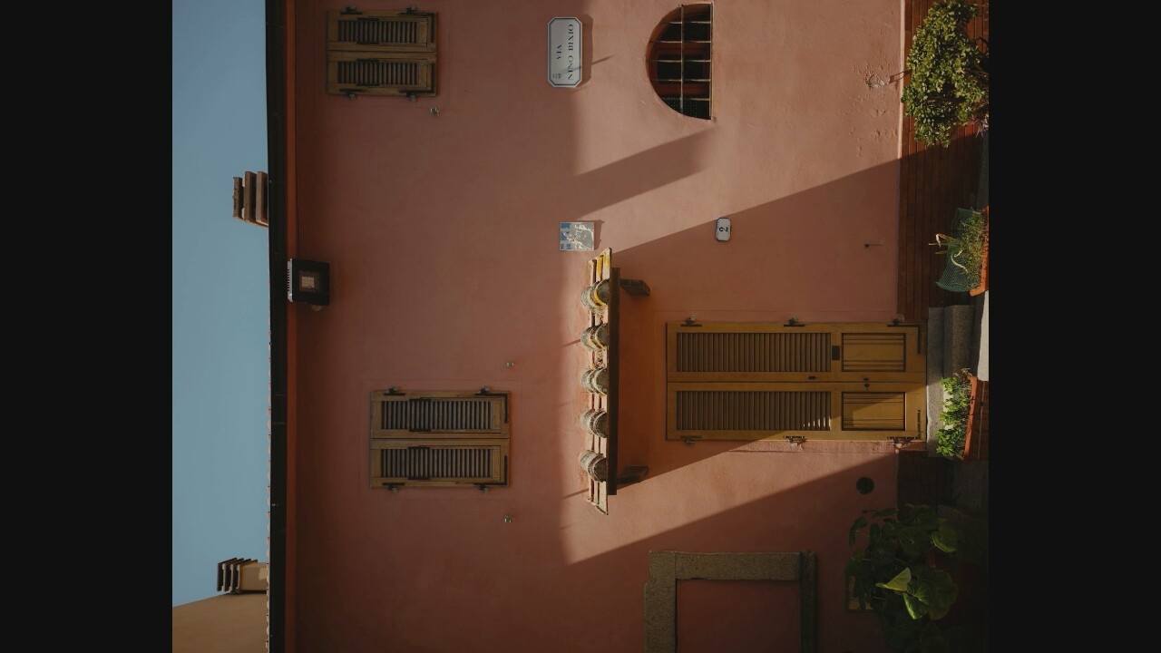 A full shot captures a pink, two-story building with an Italian street sign reading "VIA NINO BIXIO." The building is illuminated by bright sunlight, casting strong shadows. A wooden double door is central on the ground floor, flanked by steps and planters. Above the door, a small tiled awning protects the entrance. Two identical windows with wooden shutters are visible on the upper floor. To the right of the upper windows, a semicircular window with bars is set into the wall. A small blue and white tiled artwork hangs on the wall above the awning. The sky is a clear, pale blue. The bottom of the image shows plants in pots and a portion of the street.