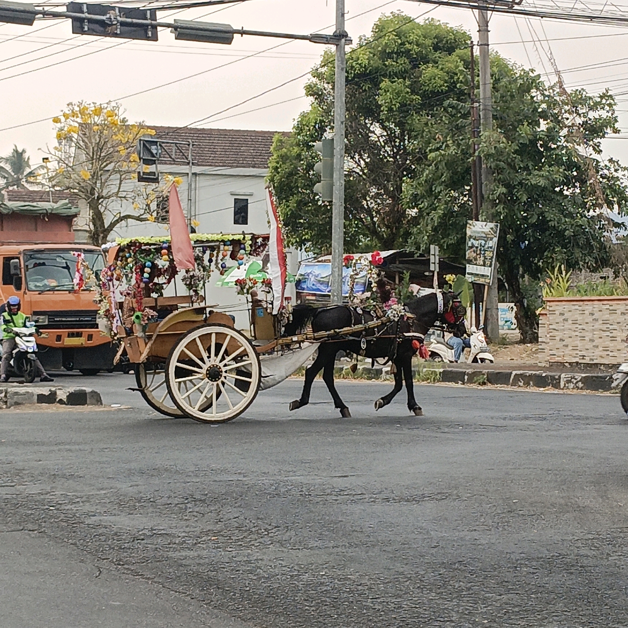 In Salatiga, Indonesia, a horse-drawn carriage adorned with colorful decorations and flags is moving across an asphalt road. The carriage has large spoked wheels and is pulled by a dark-colored horse. In the foreground, the horse's legs are in motion. To the left of the carriage, an orange truck is parked, and a person on a white motorcycle wearing a neon yellow vest and a blue helmet is stopped near the truck. The background features trees, buildings with tiled roofs, a traffic light, and overhead power lines. The sky is overcast.