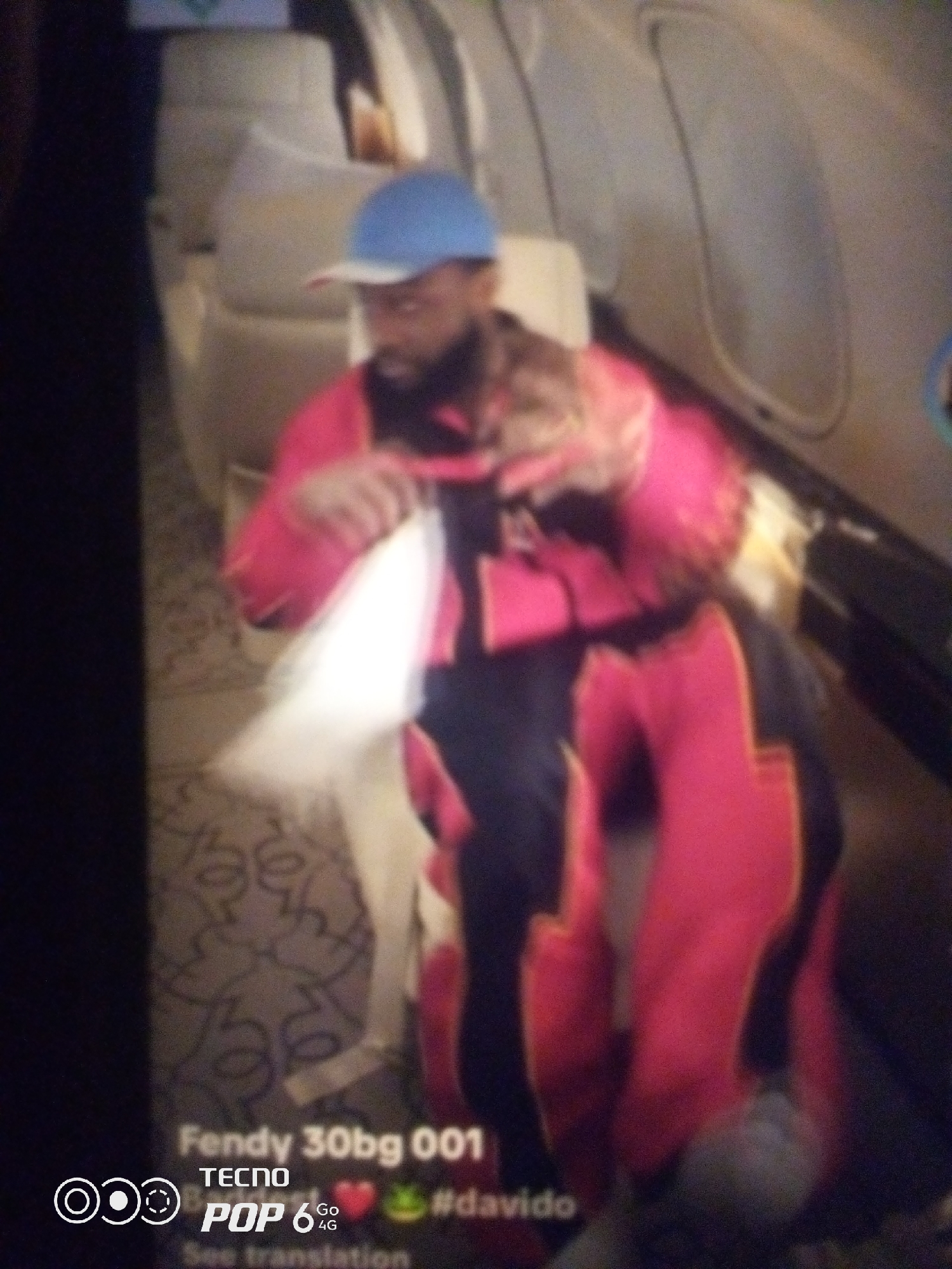 The image shows a person wearing a blue cap and a pink and black outfit, seated inside what appears to be an aircraft cabin. The person is holding a white object in their hands. The background shows the interior of the aircraft, with seats and windows visible. Text overlays on the image include "Fendy 30bg 001," "TECNO POP 6 4G," and "#davido." The image does not provide any information about the city of Howayi, Nigeria.