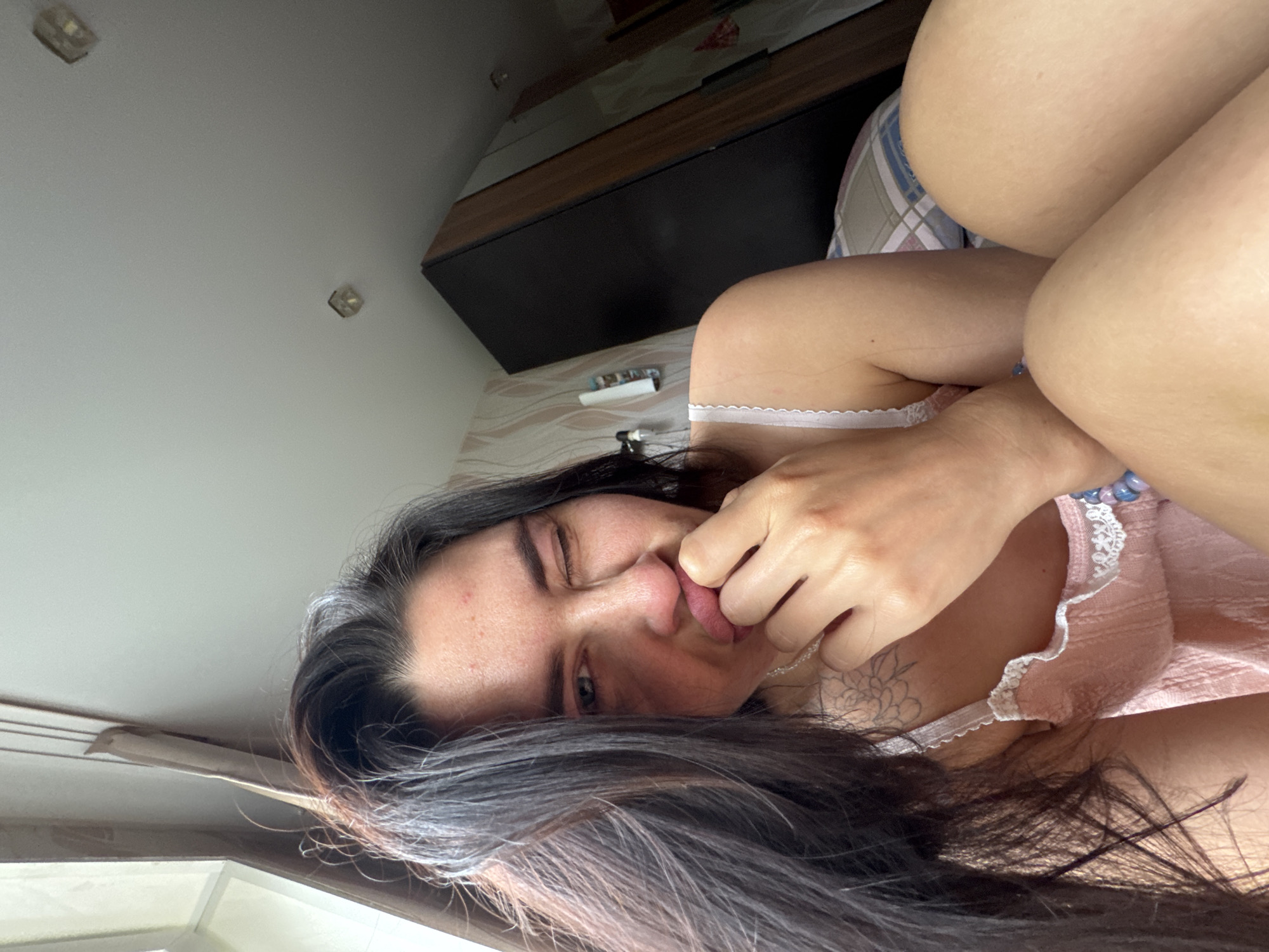 The media file contains a close-up, overhead shot of a young woman lying on her side, with her face turned towards the camera. She has long, dark hair that cascades around her head and shoulders. Her left eye is winked, and her right eye is looking towards the camera. Her right hand is touching her mouth, and her tongue is slightly extended. She appears to be wearing a pink nightgown with white lace trim. The background suggests an indoor setting, possibly a bedroom, with a dresser and a wall with a patterned wallpaper visible. The lighting indicates it could be daytime. The overall mood seems playful and candid. There is no visible text in the image.