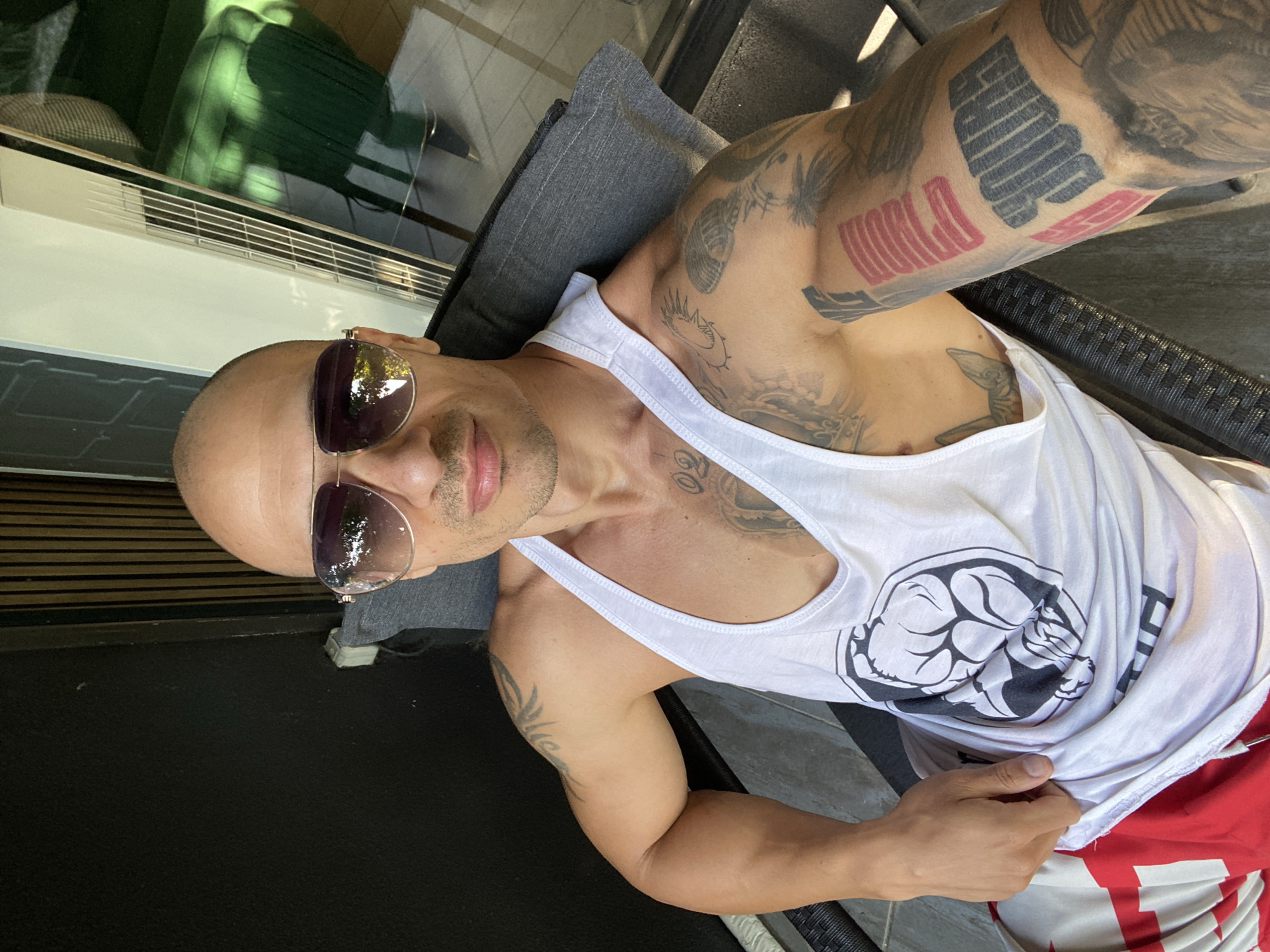 A bald man with sunglasses and tattoos is sitting in a chair outdoors. He is wearing a white tank top with a graphic design on it and red and white patterned shorts. The background shows a wooden wall and a window reflecting trees and a green chair. The location is not specified in the city of Pleven, Bulgaria.