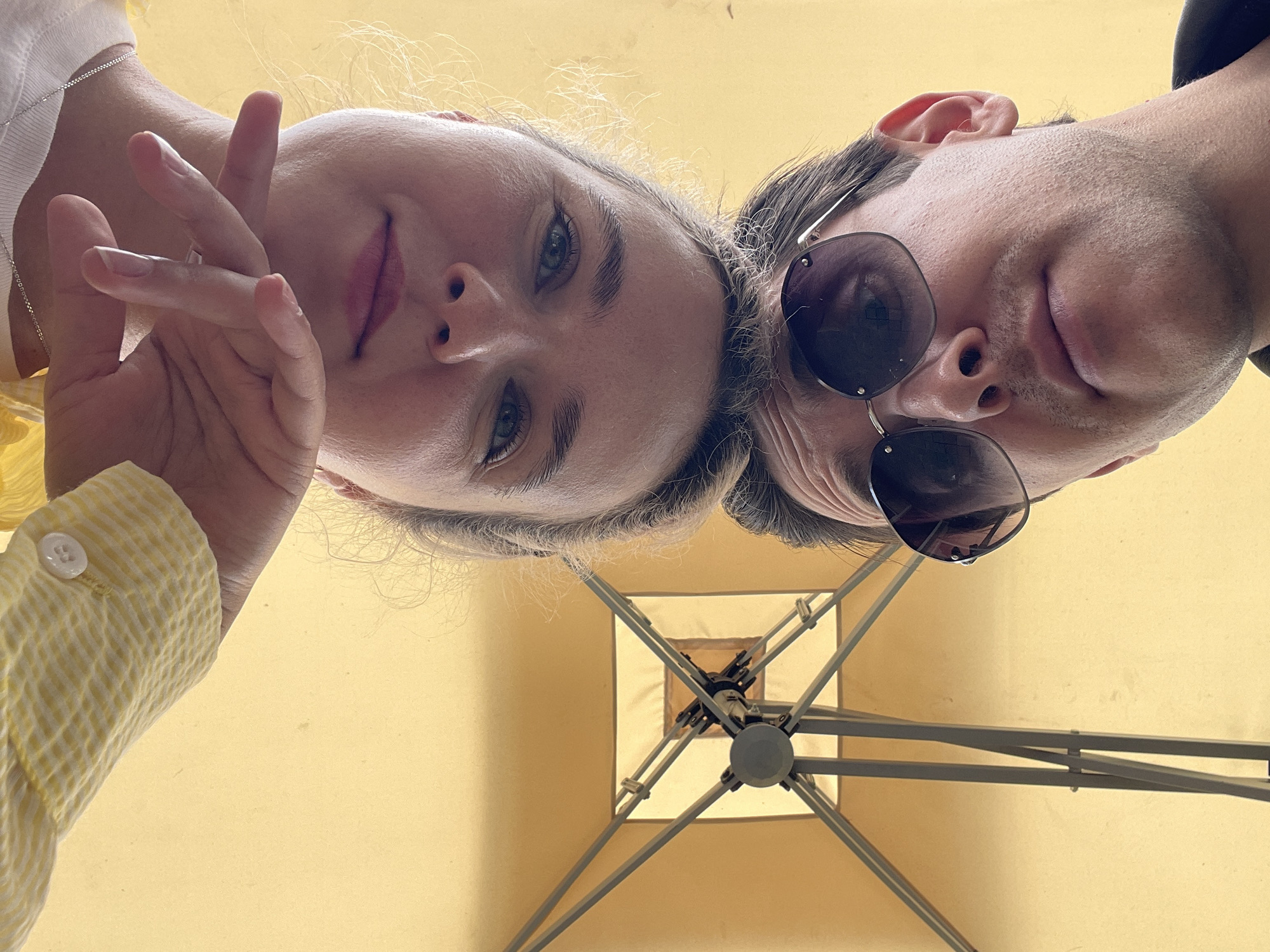 A close-up, upside-down shot captures two individuals from a low vantage point, likely under a beige umbrella. The woman, on the left, is making a hand gesture. The man, on the right, is wearing sunglasses. They are under a beige umbrella. The location appears to be Da Lat, Vietnam.