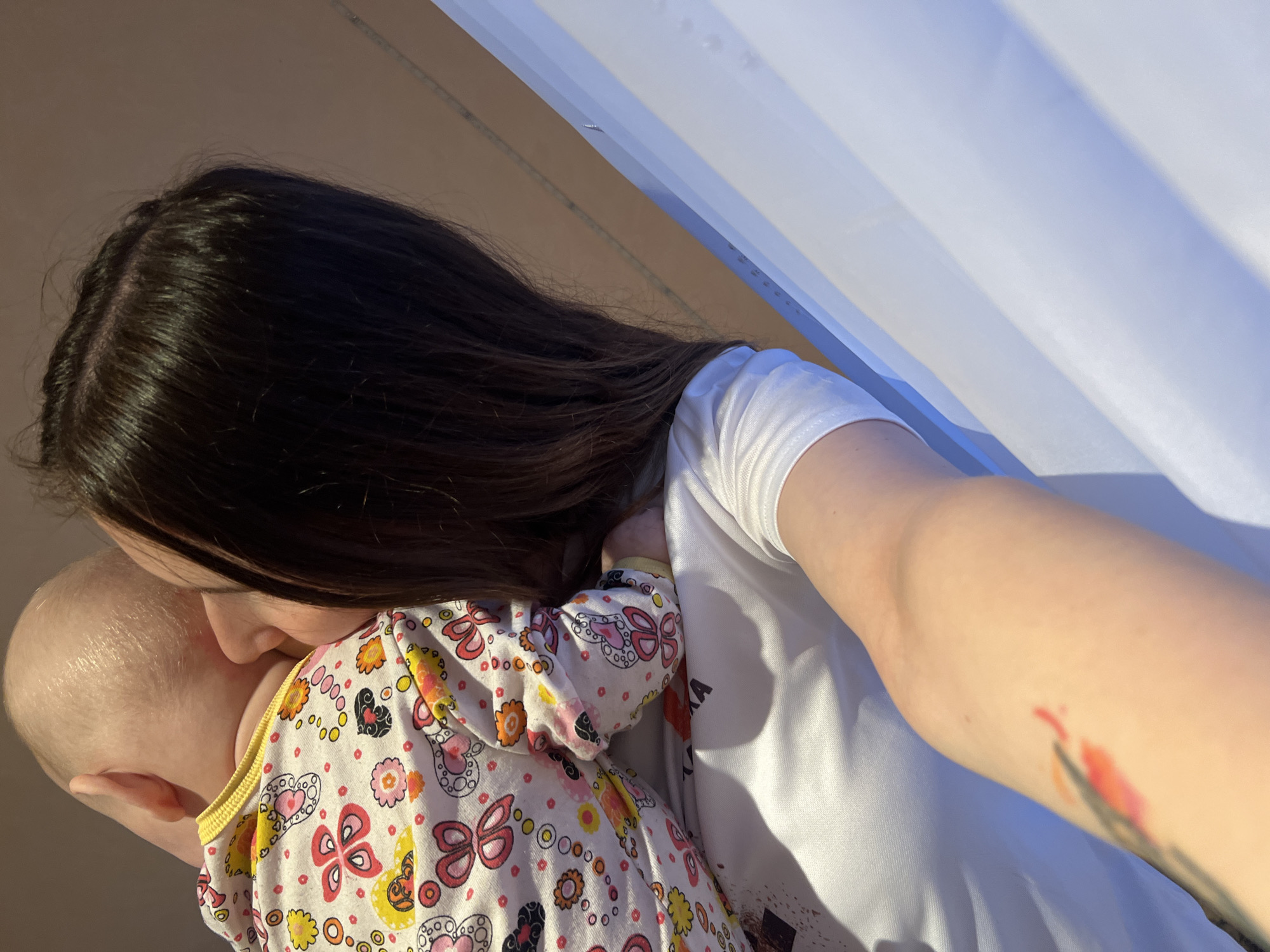 A woman is holding a baby. The woman has long, dark brown hair and is wearing a white t-shirt. The baby is fair-skinned and wearing a colorful patterned onesie. The woman is nuzzling the baby's head. The background includes a light-colored wall or surface. The location cannot be determined from the image, and there is no indication it is in Votkinsk, Russia.
