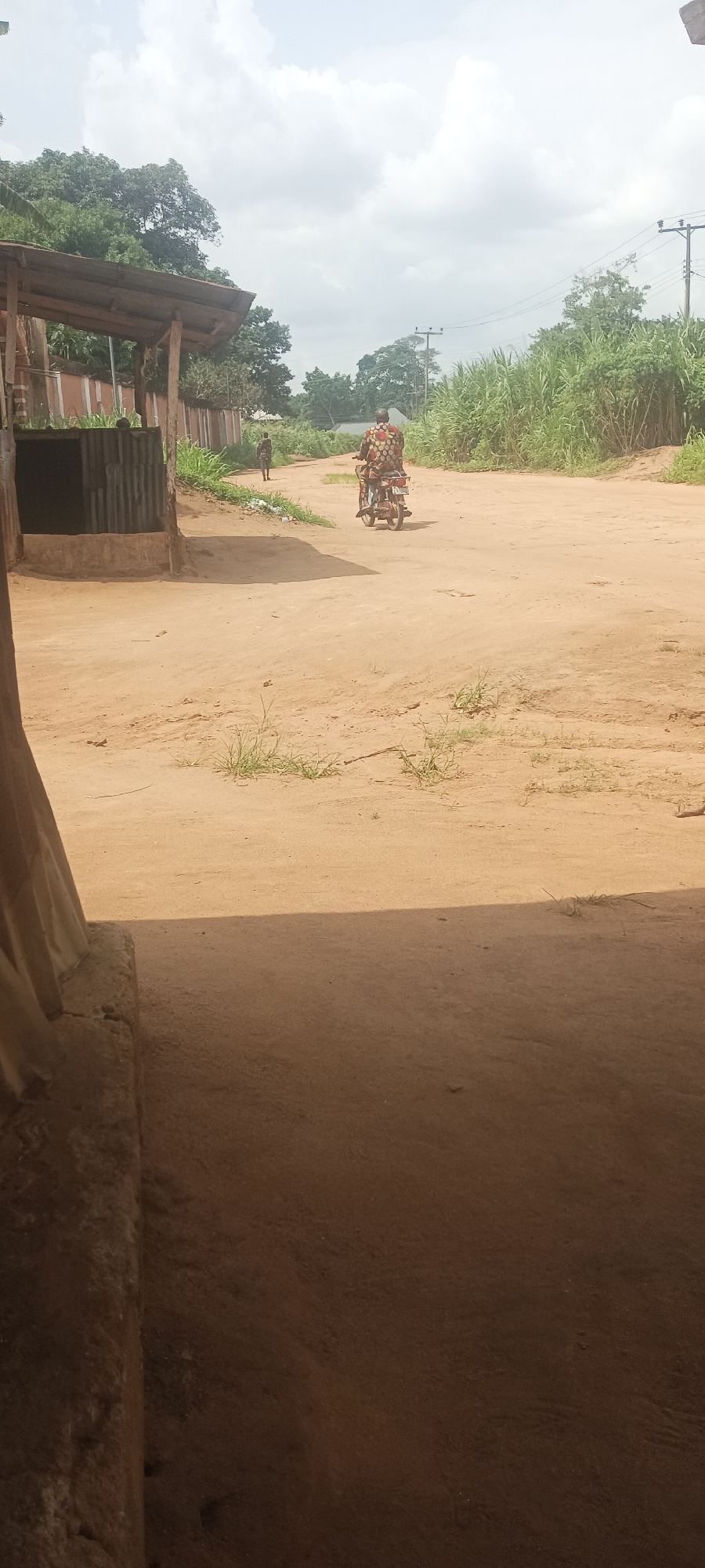 The image shows a dusty dirt road in Amauzu, Nigeria. A person on a motorcycle, wearing colorful clothing, is driving away from the camera down the road. In the distance, another person walks in the same direction. The road is flanked by vegetation and structures, including a covered area on the left and power lines in the background. The foreground is dark, suggesting the image was taken from inside a building or covered space.