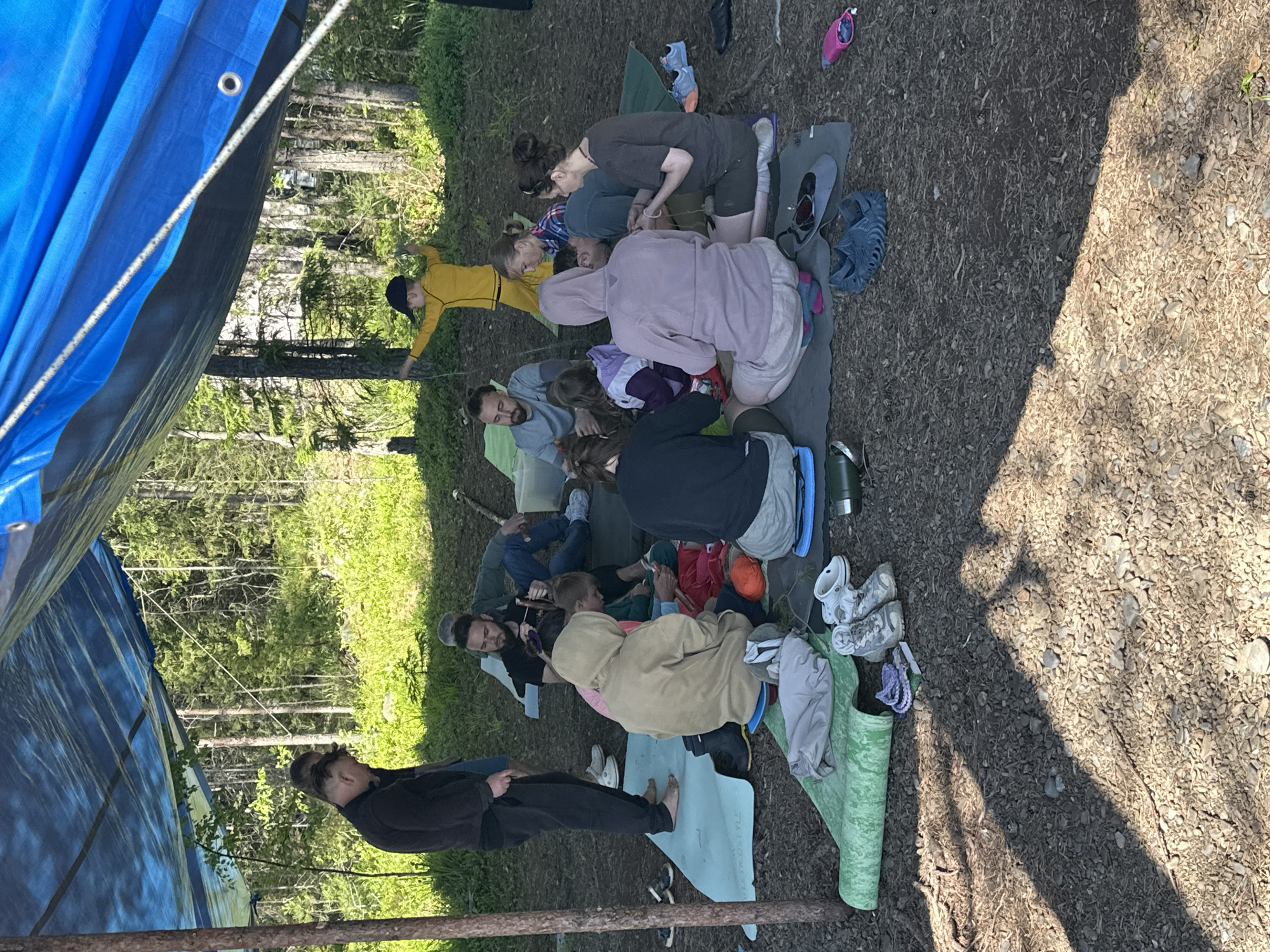 A group of people are gathered outdoors under a large blue tarp, which is supported by wooden poles. The setting appears to be a forest or wooded area, with trees and green foliage visible in the background. The ground is covered with dirt and small stones.
Several individuals are sitting or kneeling on mats or blankets on the ground, engaged in various activities. Some appear to be talking or interacting with each other. One person in a yellow tracksuit is standing and holding onto a rope or string. Children are also present in the group.
Scattered around the area are various objects including yoga mats, shoes, a water bottle, and what appear to be bags or backpacks. The overall atmosphere seems casual and relaxed, suggesting an outdoor gathering or activity.
Regarding the specific location in Soanlakhti, Russia, this image depicts an outdoor activity, likely a recreational gathering. The environment is characterized by natural elements like trees and undergrowth, which are common in the rural or forested outskirts of a city. Without more specific visual cues or landmarks, it is not possible to pinpoint a precise location within Soanlakhti. The scene suggests a temporary setup, possibly for a picnic, camping activity, or an outdoor class.