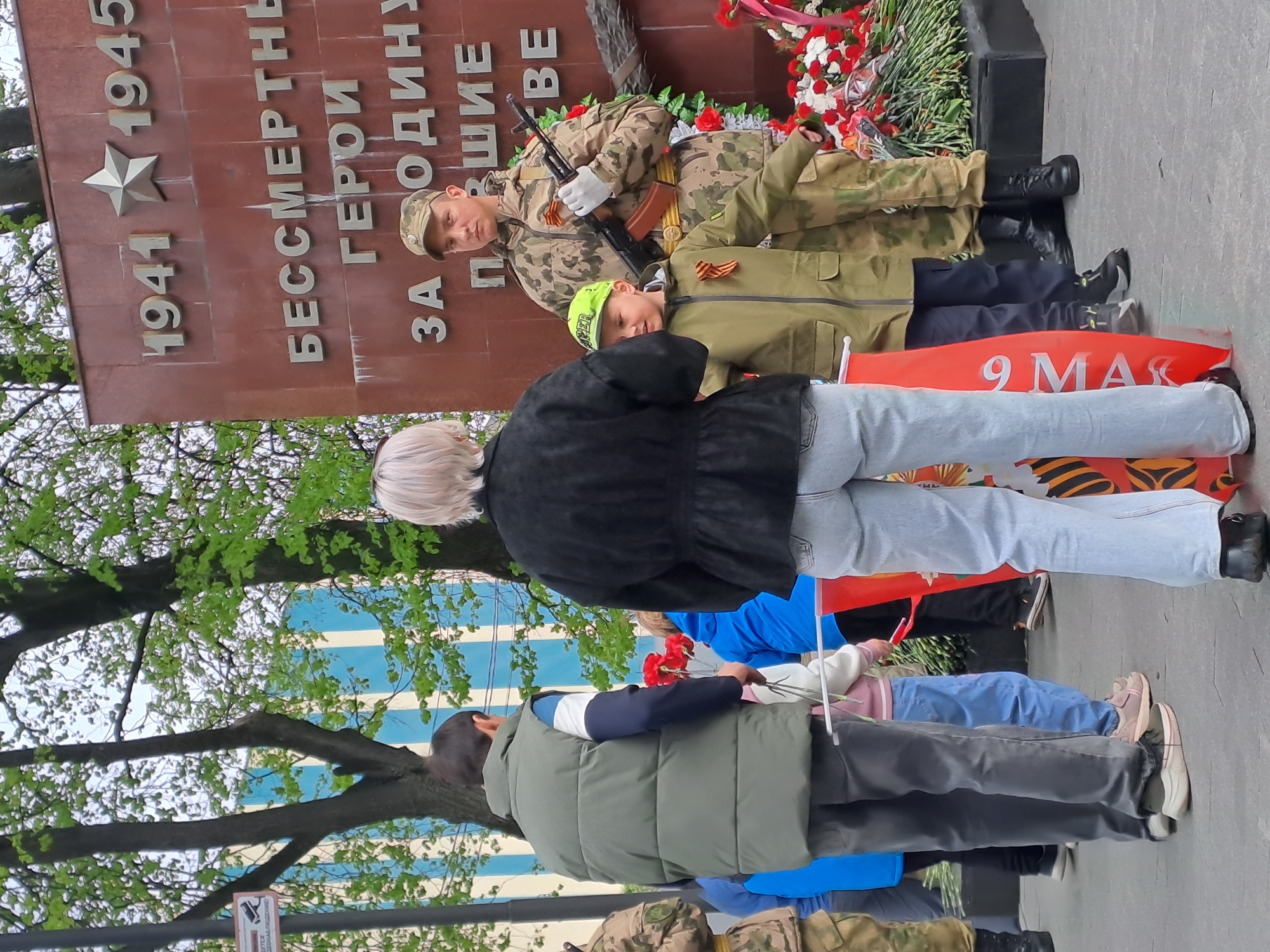 The image shows a group of people laying flowers at a memorial. The memorial is inscribed with the dates 1941-1945 and text in Russian. A soldier in camouflage uniform, holding a rifle, stands next to a child also wearing military-style clothing. The event is taking place in Elektrostal, Russia, as the memorial commemorates the Great Patriotic War.