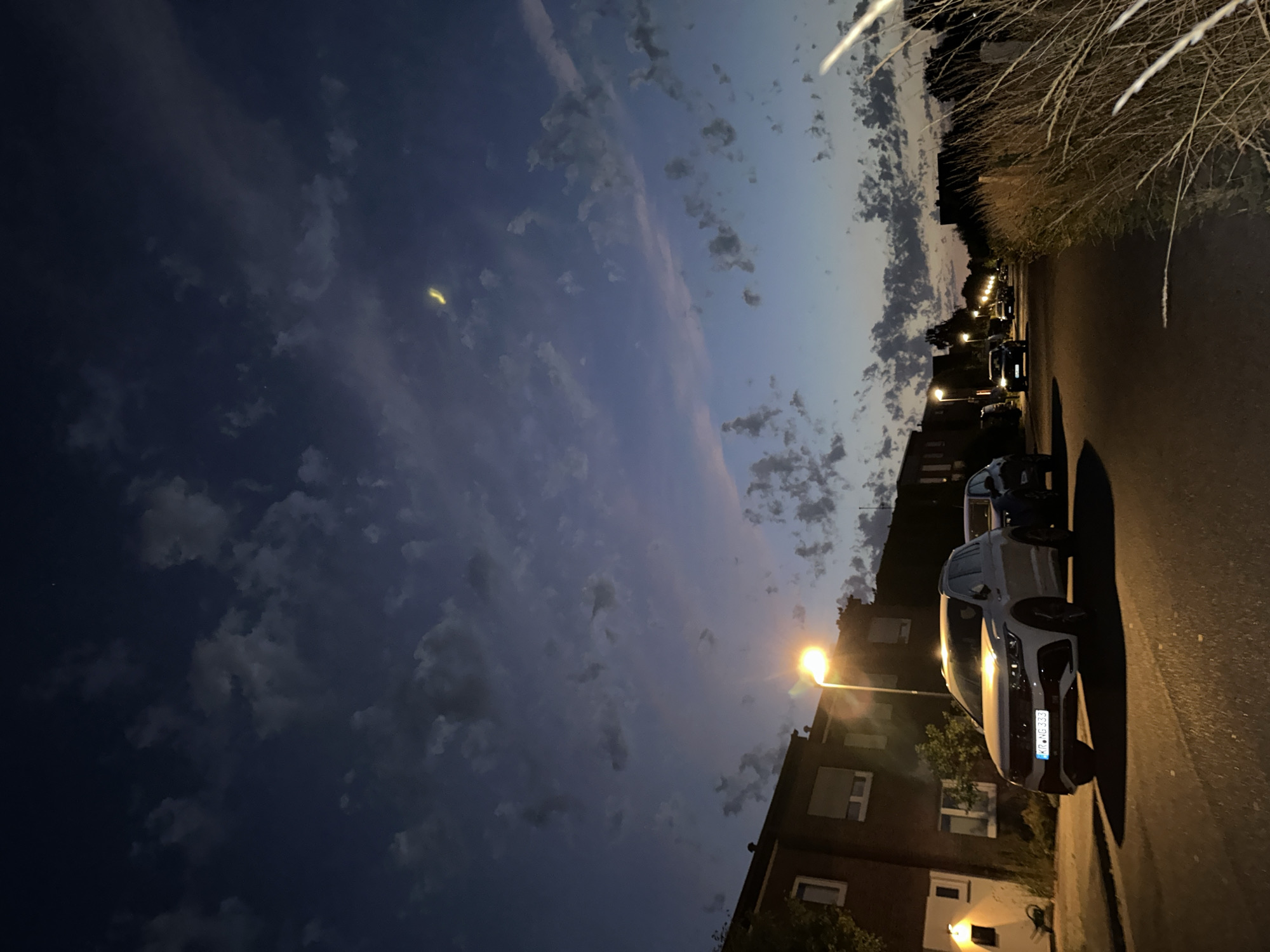 The image depicts a suburban street scene at dusk in Krefeld, Germany. The sky is a deep blue with scattered, wispy clouds, and a faint crescent moon is visible. Streetlights illuminate the scene, casting a warm glow on the asphalt road.
In the foreground, a white SUV is parked on the side of the road, facing the camera. Its license plate is visible and reads "KR-NG 333." To the right of the SUV, another car is partially visible. Further down the street, several other cars are driving or parked, and streetlights are spaced along the road.
Residential buildings, characterized by brick facades and windows, line the left side of the street. Tall grass and vegetation are present on the right side of the road, bordering a fence. The overall impression is a quiet, residential area experiencing the transition from day to night.