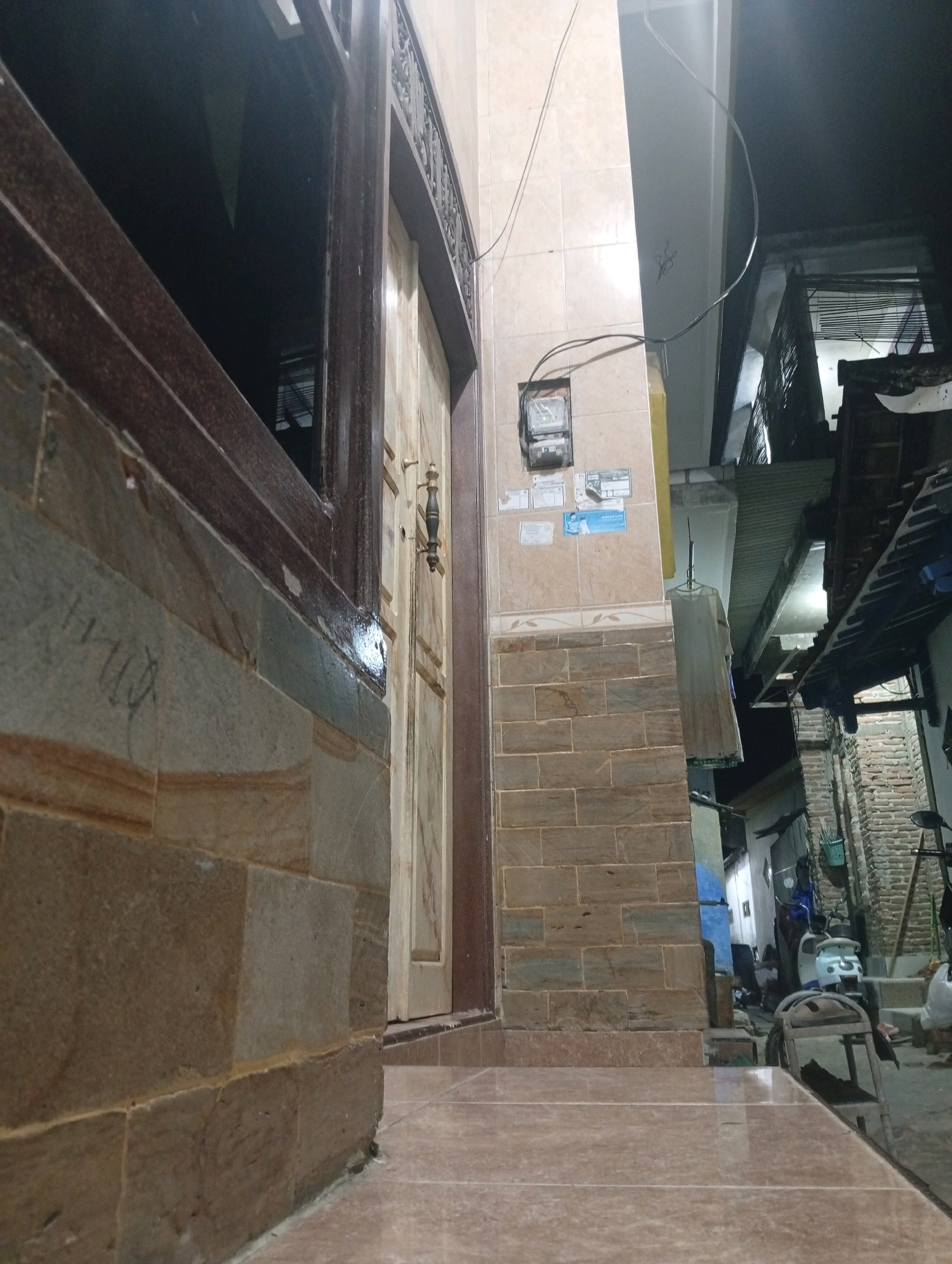 A low-angle, outdoor shot captures the exterior of a building at night, likely in Banowan, Indonesia. The foreground is dominated by a tiled walkway and a stone-clad wall. To the left, a dark window with a wooden frame reflects the dim ambient light. Adjacent to this is a wooden door with a decorative handle.
The middle ground features a column with tiled surfaces and a mounted electricity meter. Several small stickers are affixed to the tiles. To the right of the column, a white dress hangs on a hanger from a thin wire. Further back, the scene opens up to a narrow alleyway or street.
In the background, more buildings are visible, constructed with brick and corrugated metal roofing. A light-colored scooter is parked in the alley, and a person is seated on a metal chair. Another individual is partially visible behind a dark structure. The overall impression is of a residential area, possibly an urban or semi-urban setting, illuminated by artificial light.