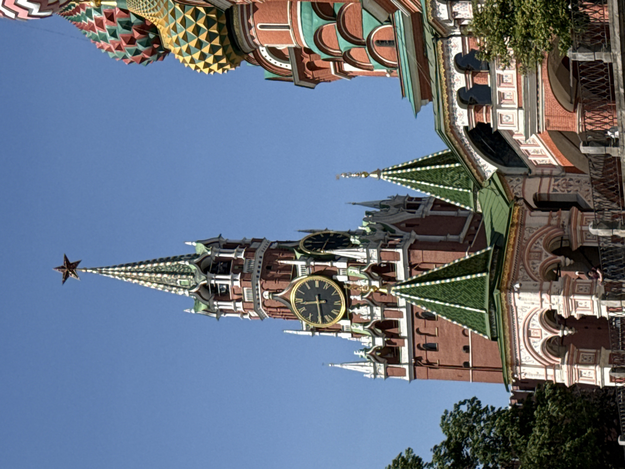 The image depicts the Moscow Kremlin in Russia. The main subject is the Spasskaya Tower, distinguished by its prominent clock and red star on top. The sky is clear and blue. The building's architecture features red brickwork, green roofs, and decorative elements. Part of St. Basil's Cathedral is also visible, recognizable by its colorful, patterned domes. Trees are visible at the bottom right corner.