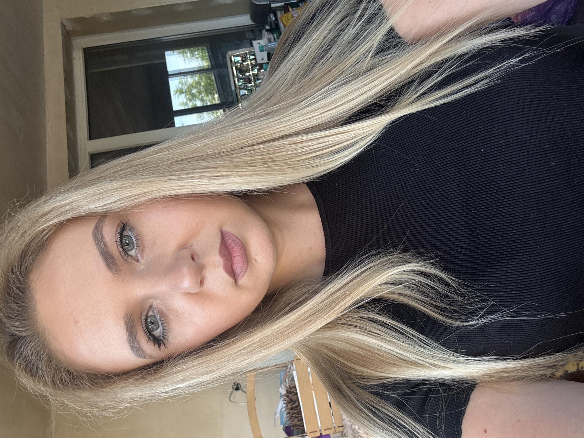 A close-up, head-and-shoulders selfie of a young Caucasian woman with long, blonde hair styled straight. She is wearing a black, ribbed, short-sleeved shirt. Her eyes are light blue and she has on makeup, including mascara and lipstick. The background appears to be outdoors, with a window or glass door visible behind her, reflecting trees and the sky. To her left, a wooden crate filled with what appear to be pinecones is partially visible, along with a lamp and some other objects. The lighting suggests it is daytime.