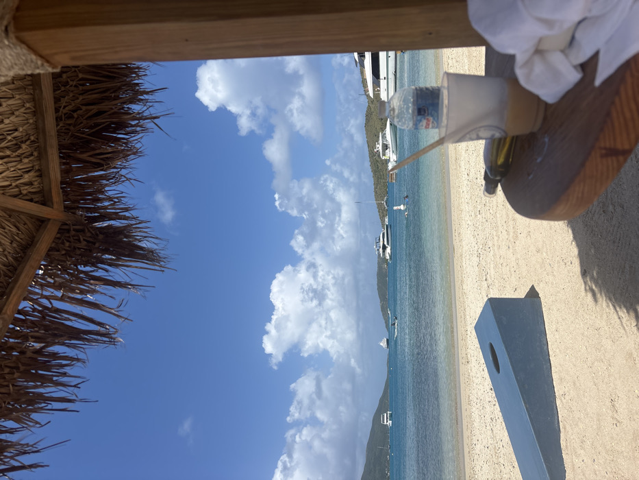 The image depicts a sunny daytime scene on a sandy beach in Spanish Town, British Virgin Islands, viewed from beneath a thatched roof structure with a wooden support post visible on the right.
In the immediate foreground on the right, a circular wooden table displays a clear plastic cup containing a light-colored beverage and a wooden stir stick. A "Crystal Geyser" plastic water bottle rests inside the cup. Adjacent to the cup, a dark-colored bottle lies horizontally, and a crumpled white cloth or towel is present. The thatched roof, composed of dried fronds, overhangs the top portion of the frame, casting a shadow over the foreground.
The mid-ground features a light-colored sandy beach. A blue-painted cornhole board is positioned on the sand, angled towards the water. Beyond the beach, calm, clear turquoise water extends across the frame. Multiple vessels are anchored or moored in the bay, including at least five large white yachts, several smaller boats, and sailboats. A dark-hulled vessel is situated near the center of the bay. Two individuals are present in the water: one appears to be on a white paddleboard or inflatable, while another is partially submerged nearby.
The background consists of green, vegetated hills forming distant shores under a clear blue sky dotted with scattered white cumulus clouds.