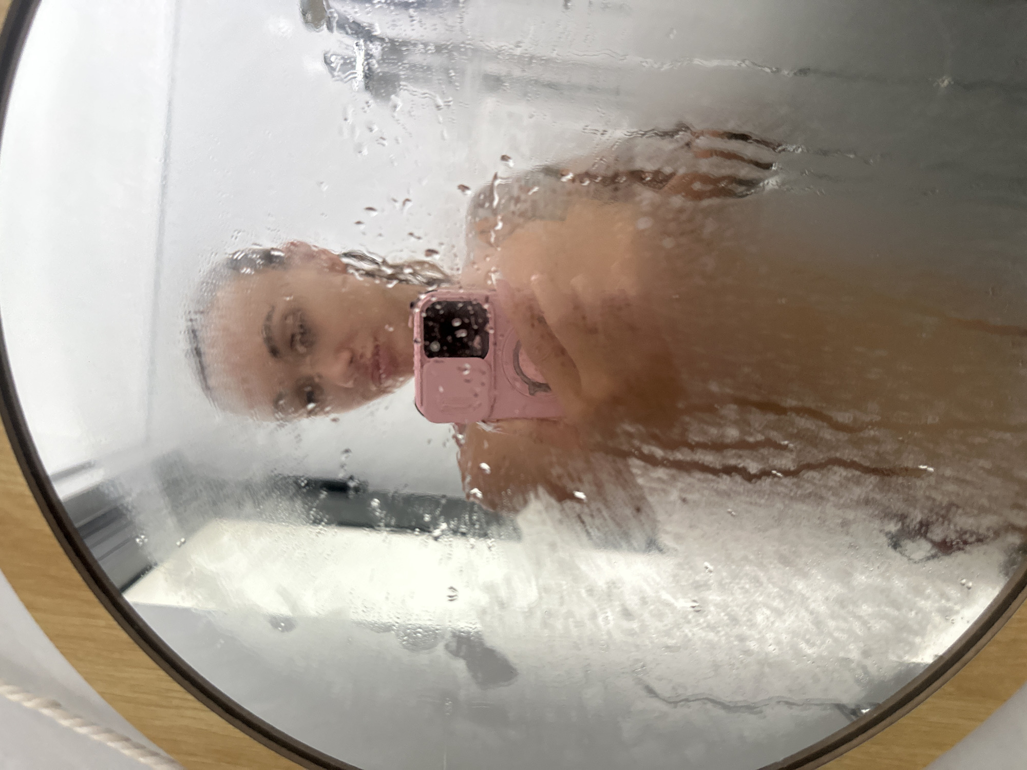 A selfie is captured in a round, wooden-framed mirror, heavily obscured by water droplets and condensation, suggesting a steamy bathroom or shower setting. The reflection shows a person, likely female, holding a pink smartphone to take the picture. Her face, seen through the water-streaked glass, is turned slightly towards the camera with a neutral to soft expression, and her hair appears wet. The pink smartphone's camera array is visible in the reflection, partially blocking her face and torso. The background reflection, distorted by the water, shows a light-colored area, possibly a tiled wall or the interior of a shower stall. The image conveys a personal moment, possibly taken during or immediately after a shower. The implied location context for this indoor scene is Mũi Né, Vietnam. There is no visible text in the image.