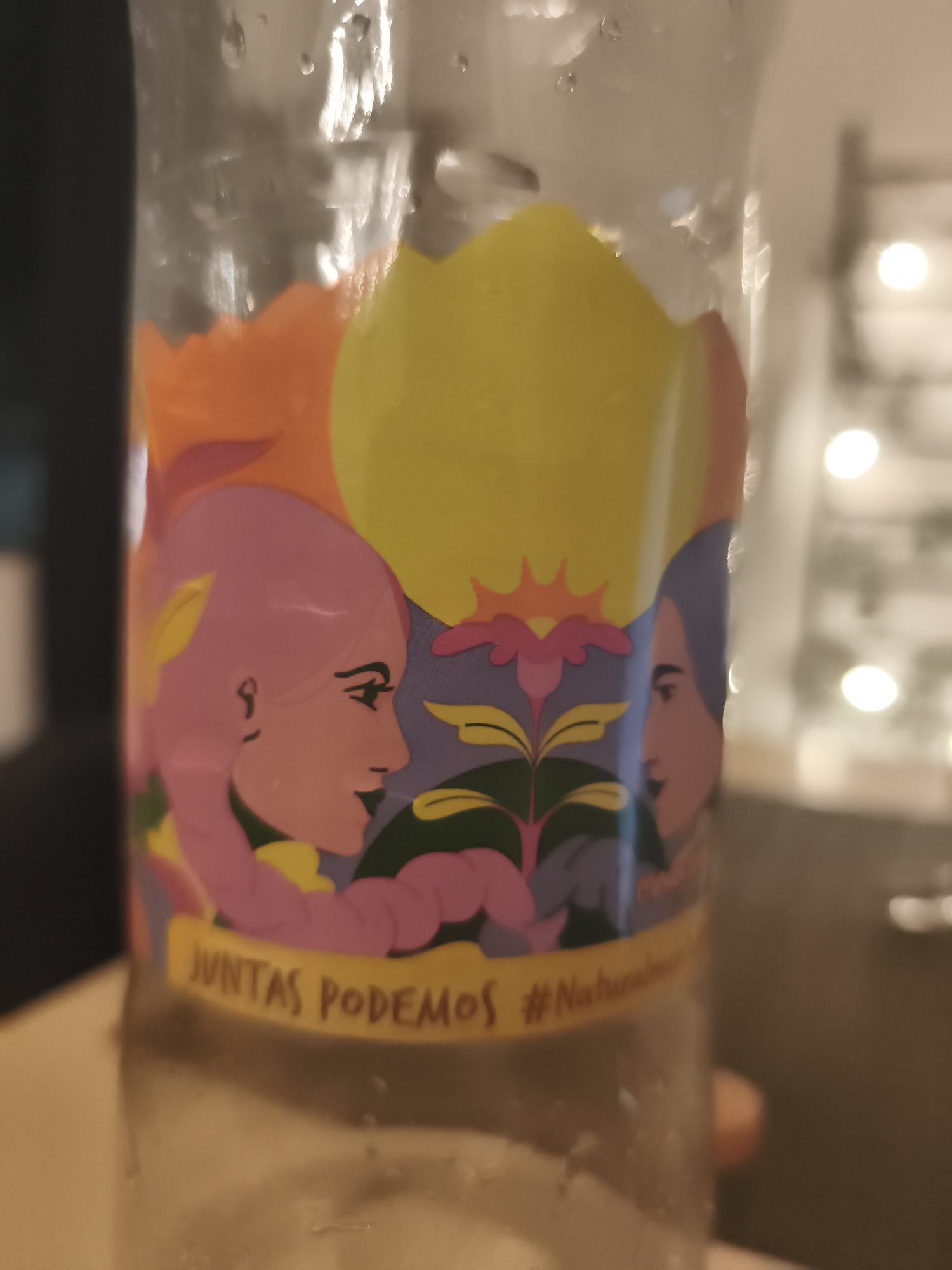 A clear plastic bottle is shown in close-up, featuring a colorful illustration. The illustration includes two stylized profile faces: one with pink hair and the other with purple hair. They are facing each other. Above them, there is an orange shape and a yellow circle, possibly representing the sun. In the center, there is a stylized plant with a purple flower and green leaves. Below the illustration, there is a yellow banner with the text "JUNTAS PODEMOS #Naturalmente". The bottle appears to be partially filled with a brownish liquid.
The background is blurry, showing a room with soft lighting. The presence of bokeh suggests the photograph was taken indoors. There are no apparent indications of Mexico City, Mexico in the image.