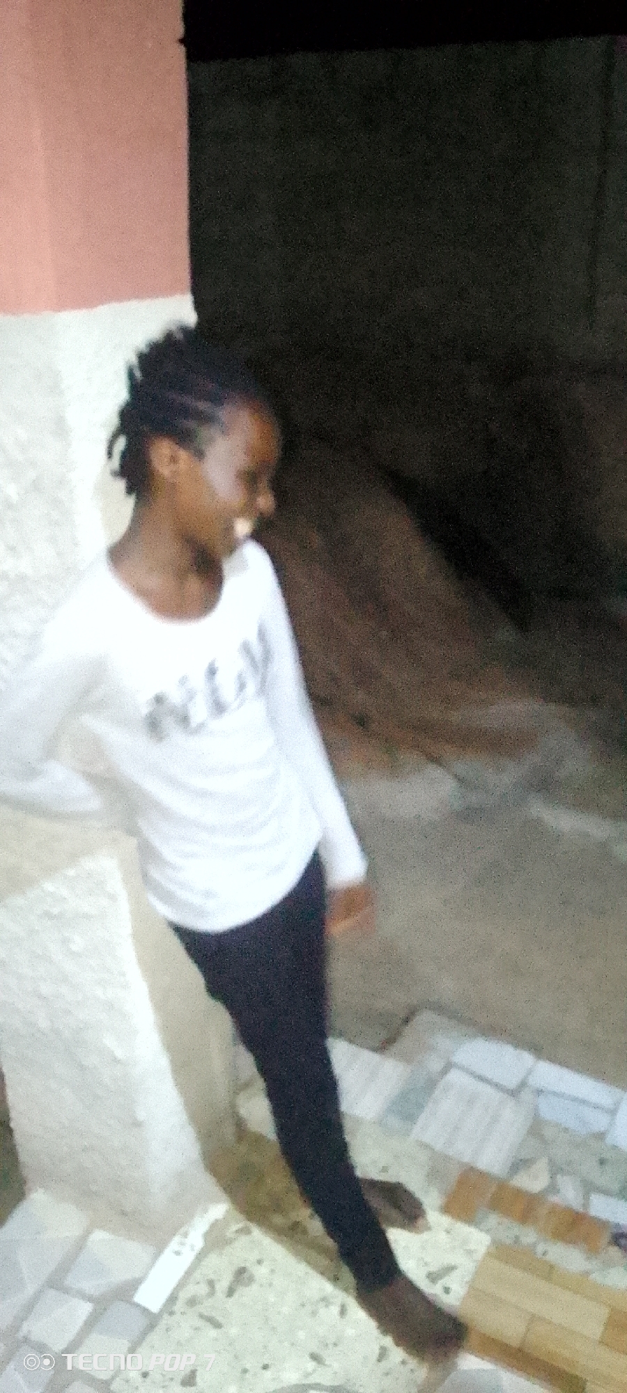A full-body, slightly blurry, vertical outdoor shot shows a young Black female standing and leaning against a light-colored wall on the left. She has braided hair and is wearing a white long-sleeved shirt with a graphic design on the front and black leggings. Her head is tilted down and to her right, with her mouth open as if she is laughing or speaking. She is standing on a tiled surface. The background consists of a dark, unlit area on the right and a reddish-pink wall on the upper left. The bottom of the image shows a portion of the TECNO POP 7 watermark.