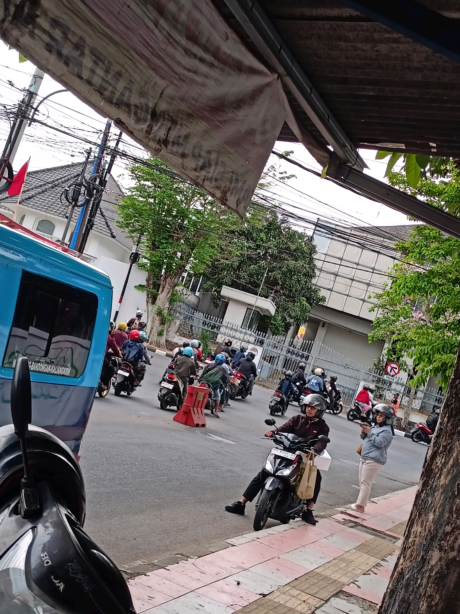 The image depicts a street scene in the city of Magelang, Indonesia. A convoy of motorcycles navigates a paved road lined with sidewalks and buildings. The vehicles are of various makes and models, and the riders are predominantly wearing helmets for safety. Some riders carry bags, suggesting they may be on errands or making deliveries. A blue bus is partially visible on the left side of the frame, indicating public transportation is also present. The environment appears to be a typical urban setting with overhead power lines, trees, and a building with a distinctive facade. The road has markings and a red traffic cone is visible, hinting at potential traffic management or construction. The overall atmosphere is that of a busy street with ongoing activity.