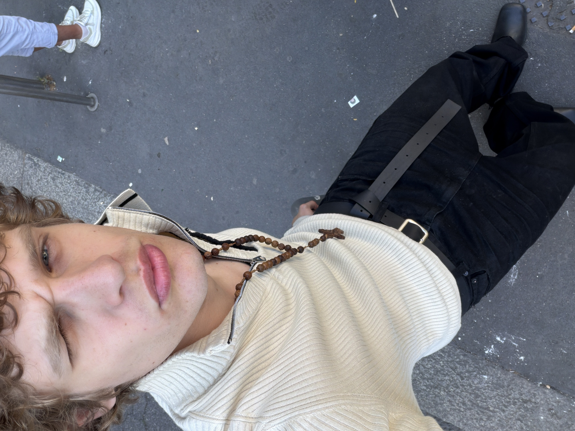 A person with curly blond hair is photographed from above, with their head angled to the left. They are wearing a light-colored ribbed zip-up sweater and a beaded necklace with a cross. Their lower body is clad in black jeans with a black leather belt and black shoes. In the upper left corner of the frame, a portion of another person wearing white sneakers and light blue pants is visible. The scene is set on a gray asphalt surface with scattered debris. The location is not identified as Milan, Italy.