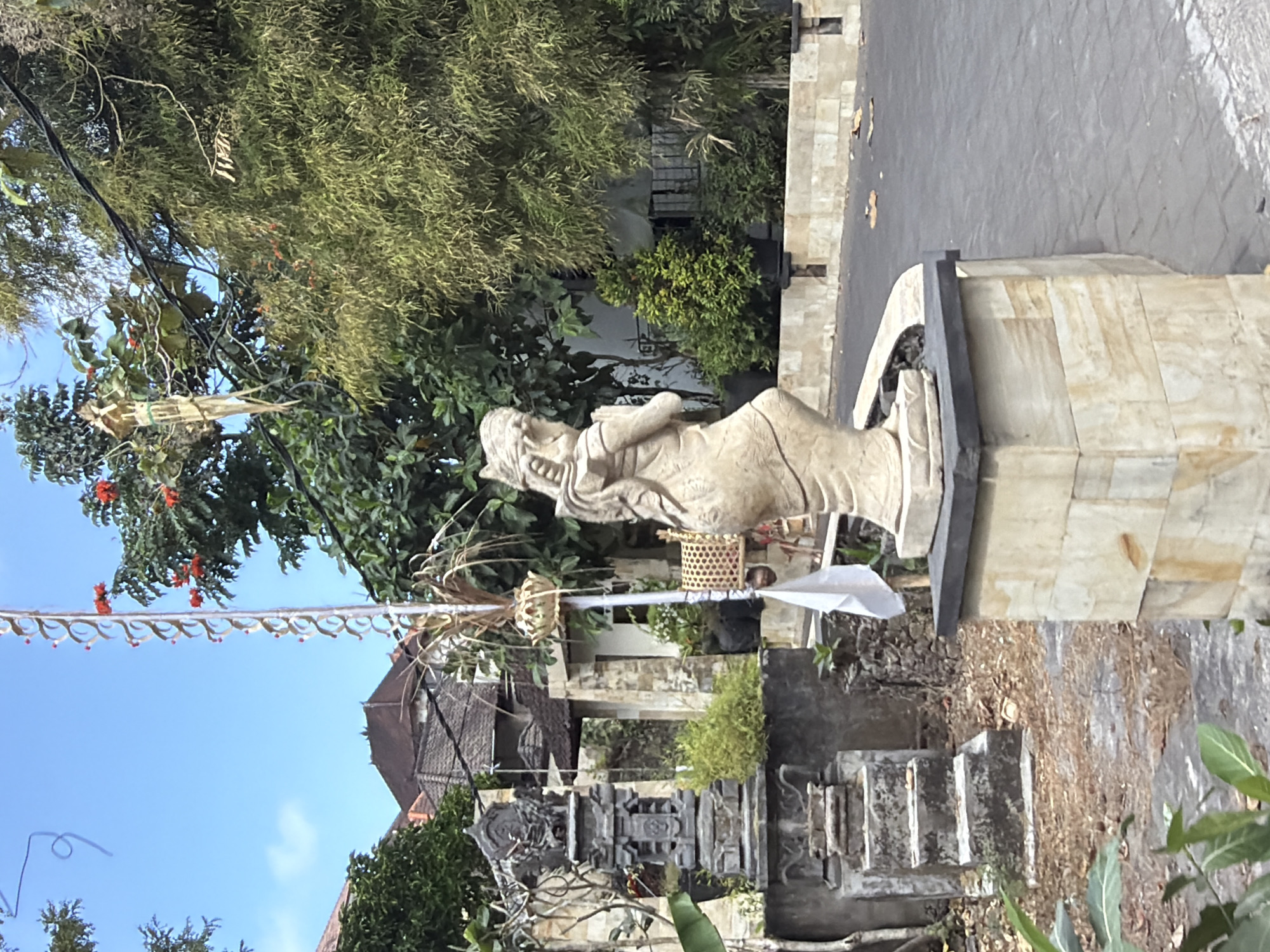 The image depicts an outdoor scene with traditional Balinese architectural elements. In the foreground, a light-colored stone statue of a woman in a prayer pose stands on a pedestal. To the left, a tiered stone structure, possibly a shrine or a monument, is visible. A white flag is attached to a pole behind the statue. The background reveals lush greenery, including trees and bamboo, and traditional Balinese houses with dark tiled roofs. The ground is paved with dark stones and some dry leaves are scattered. The sky is clear and blue. This scene is likely located in Kuta Selatan, Bali, Indonesia, a region known for its spiritual sites and unique cultural aesthetics.