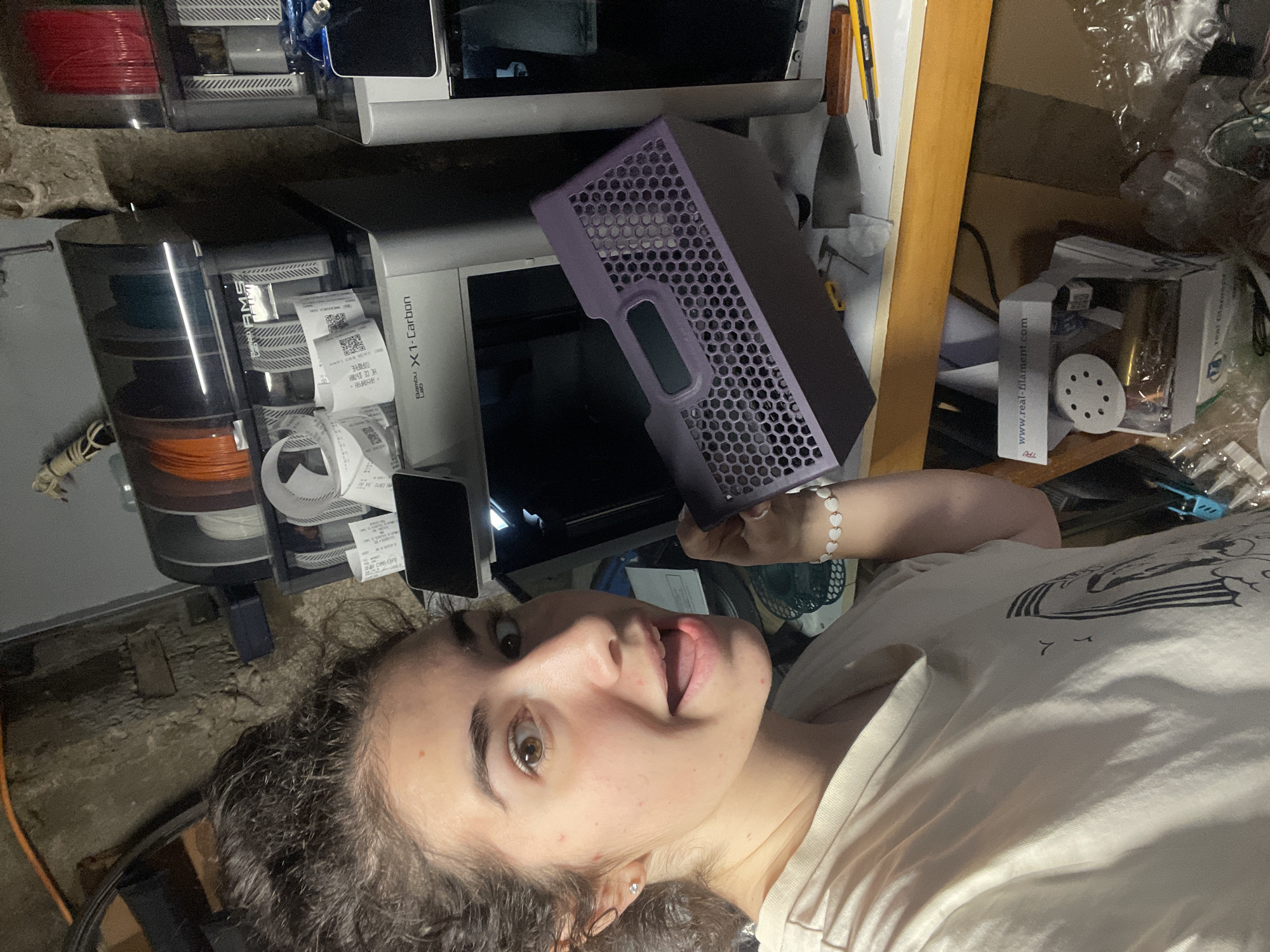 A selfie captured in a cluttered room shows a fair-skinned person with curly brown hair, wearing a white T-shirt and a bracelet with heart-shaped beads. The person is smiling with their tongue slightly sticking out. Behind them is a Bambu Lab X1-Carbon 3D printer with an AMS filament system. There's a purple, honeycomb-patterned object in the person's hand, possibly related to the printer. Various tools, filament spools, and packaging materials are scattered around, suggesting a workshop or maker space. Based on the printer, it's likely a modern, indoor setting, but there's insufficient information to place it within the city of Sofia, Bulgaria.