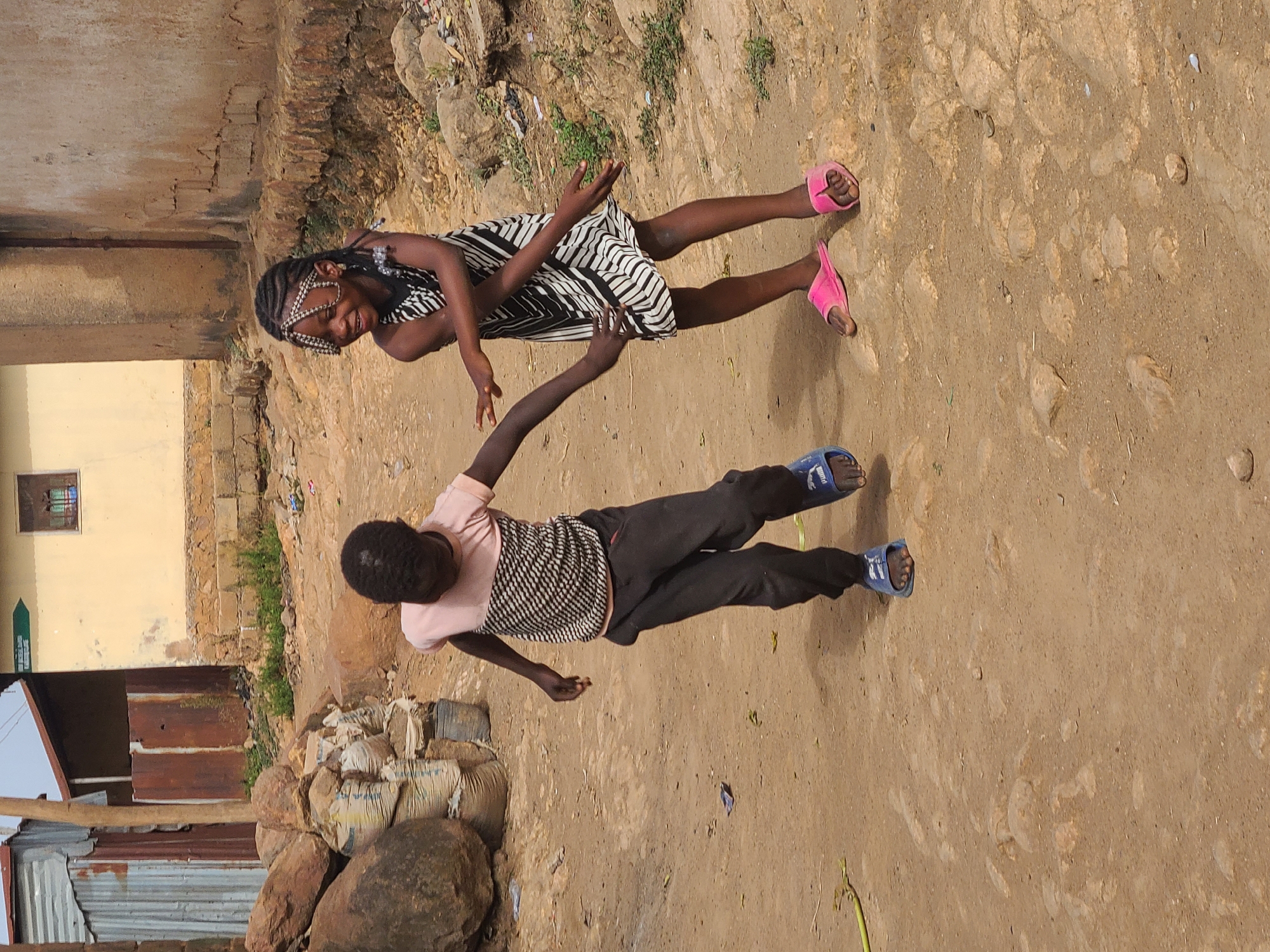 Two children stand on a dirt road, likely in an informal neighborhood of Jos, Nigeria. One wears a black and white striped dress and pink sandals. The other, facing forward, wears dark pants, a pink shirt, and blue sandals. They are interacting with their hands touching. The background features a mix of weathered buildings made of brick, concrete, and corrugated metal. A pile of stones and bags sits by one structure. The ground is dry and dusty.