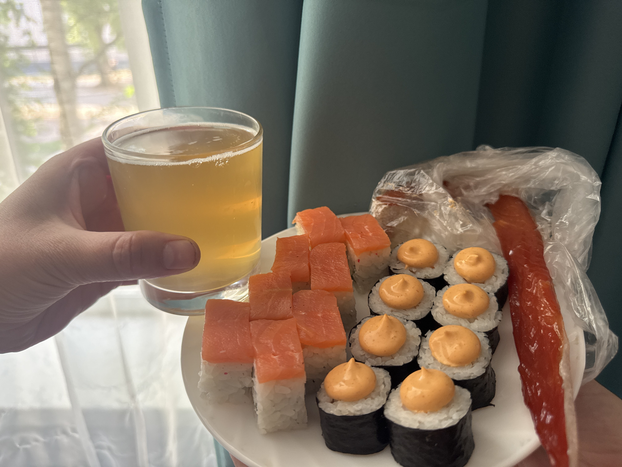 The image presents a close-up shot of a hand holding a plate of sushi and a glass of beer. The sushi appears to be a combination of salmon maki rolls and other sushi rolls topped with an orange sauce. Next to the sushi is a piece of what looks like smoked salmon in a plastic bag. The beer is light in color with some visible bubbles. The background includes a window with a sheer white curtain and a teal-colored curtain. The city is specified to be Chaikovsky, Russia, but there are no details confirming the location. The image's content is limited to food, beverages, and a blurred background with no further forensic information about the people, subjects, objects, actions, interactions, or the environment.