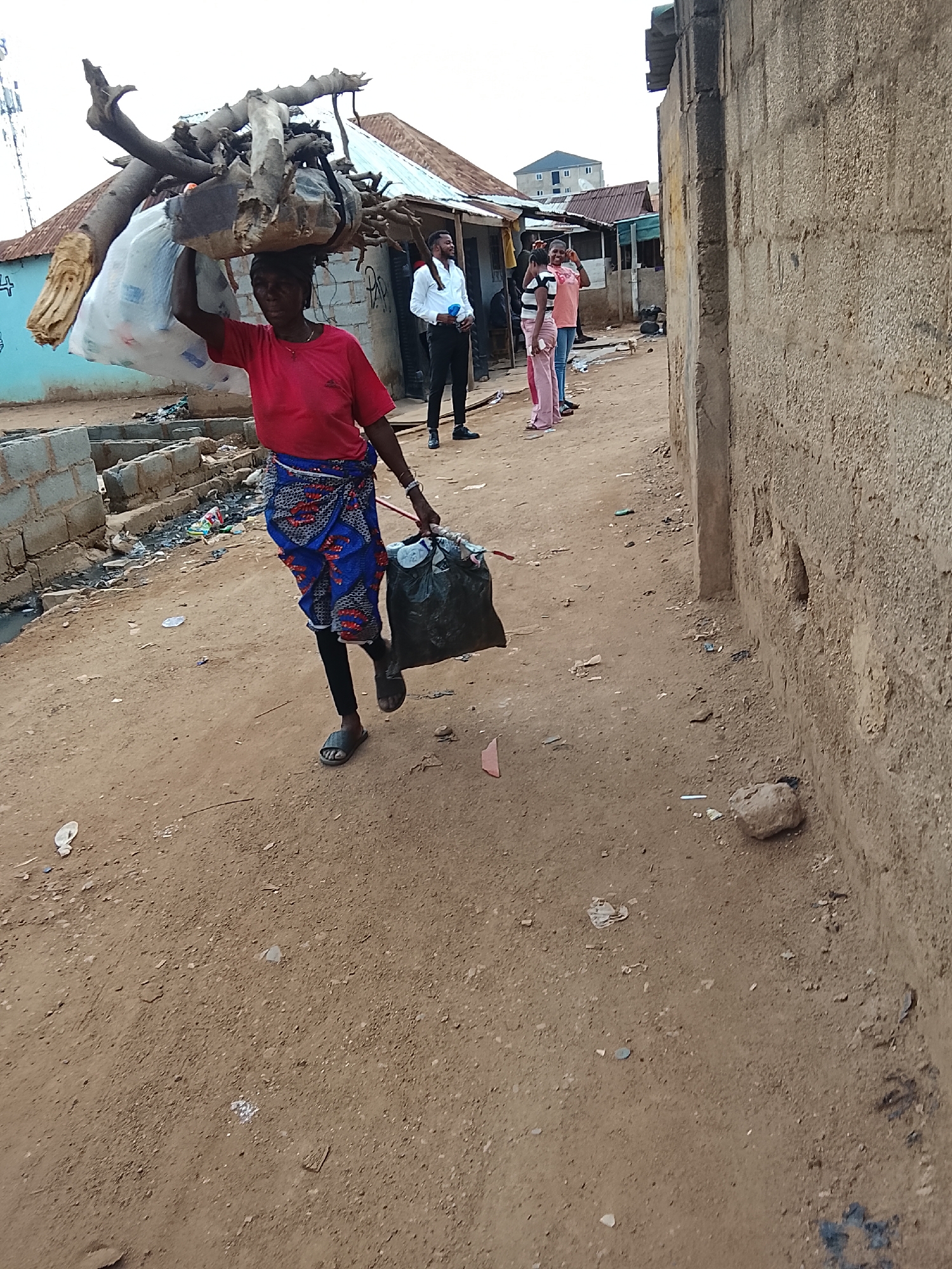 A woman is carrying a load of wood on her head, as well as a trash bag in her left hand. She is wearing a red shirt, a blue and red patterned wrap skirt, black leggings, and sandals. She is walking on a dirt road in a neighborhood in Abuja, Nigeria. In the background, there are several other people and buildings. The buildings are made of concrete blocks and have corrugated metal roofs. There is trash on the side of the road. A man in a white shirt and black pants is standing nearby. Two women in pink clothing are standing behind him. A concrete wall is in the foreground on the right.