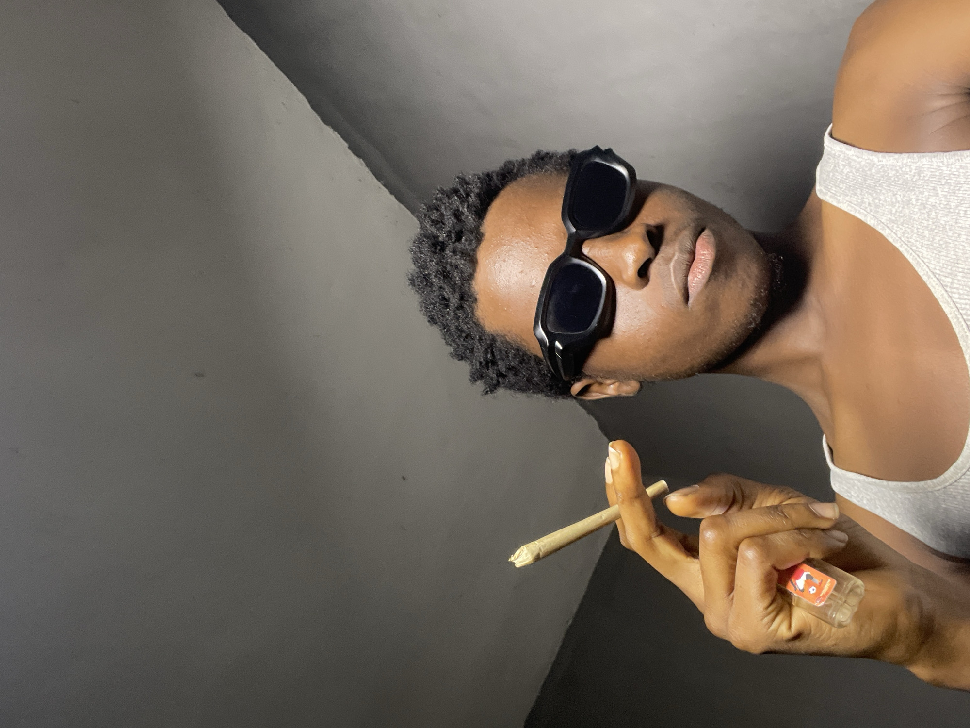 A young Black man wearing sunglasses and a white tank top is holding a cigarette and a small, clear bottle with an orange label. He is looking directly at the camera. The background is a plain, gray wall. The scene is indoors, possibly in a residential setting. There is no indication of the location being in Nkpor, Nigeria.