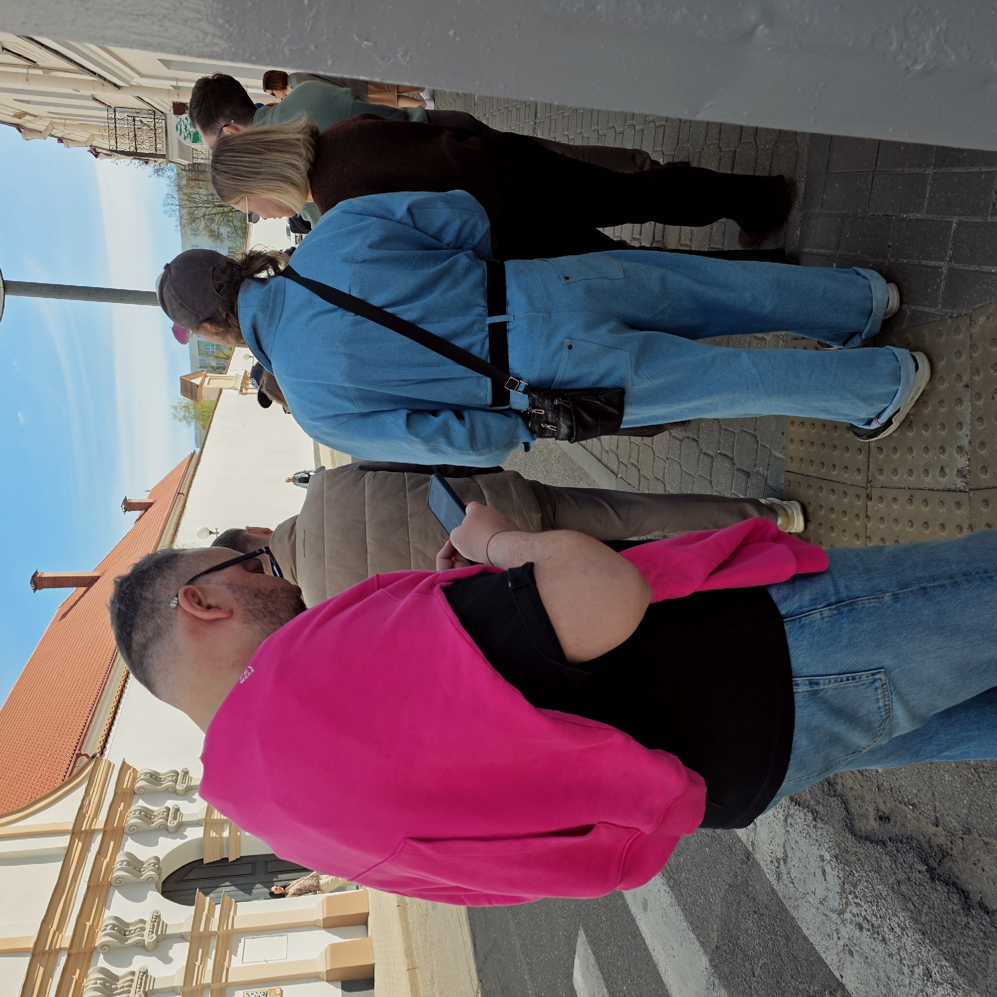The image captures a street scene in Hrodna, Belarus, with a group of people standing along a sidewalk. At the forefront of the image, a man wearing glasses and a bright pink hoodie is visible, his back partially turned. Next to him stands another individual wearing a tan jacket and holding a smartphone. Further down the line is a person in a blue denim jacket with a black cross-body bag, followed by two more people; one wearing a brown jacket and another with a green shirt. They appear to be standing in a line. The background features a building with a terracotta tile roof and a white façade with decorative architectural elements. A blue sky with some clouds is visible above the buildings. The sidewalk is paved with gray stone blocks.