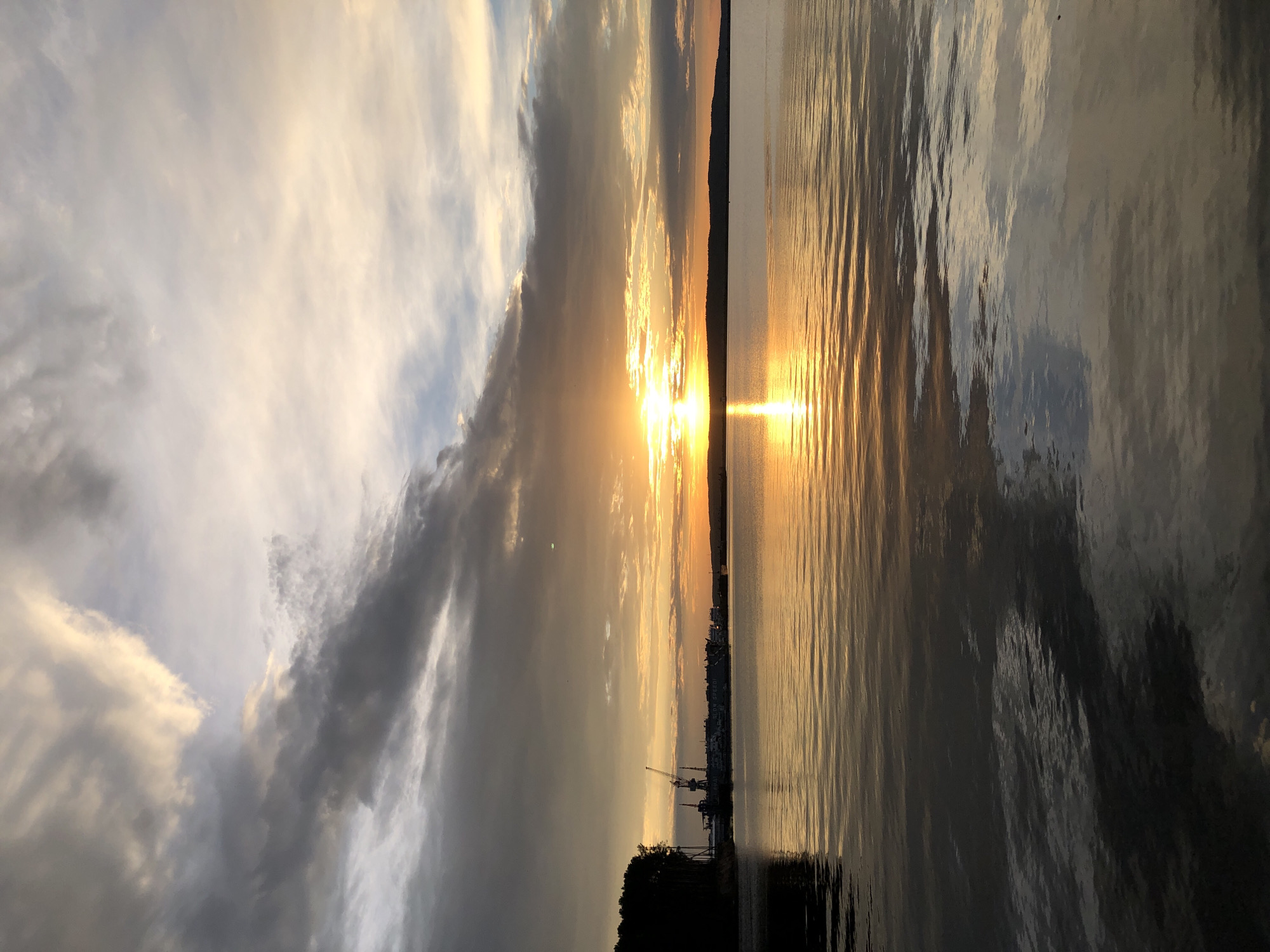 A wide shot captures a tranquil sunset over a body of water, likely a reservoir on the Kama River, near the city of Chaikovsky, Russia. The sky is a canvas of soft oranges, yellows, and grays, with dramatic clouds reflecting the fading sunlight. The sun itself, a brilliant orb, is low on the horizon, casting a golden shimmer across the rippling water.
On the left side of the frame, a dark, silhouetted line of trees marks the shoreline, behind which are industrial structures, including cranes and docked boats, suggesting a port or industrial area. The water occupies the majority of the lower half of the image, mirroring the sky and the distant landscape with gentle waves. The overall scene evokes a sense of peace and the end of the day in this industrial yet naturally beautiful setting.